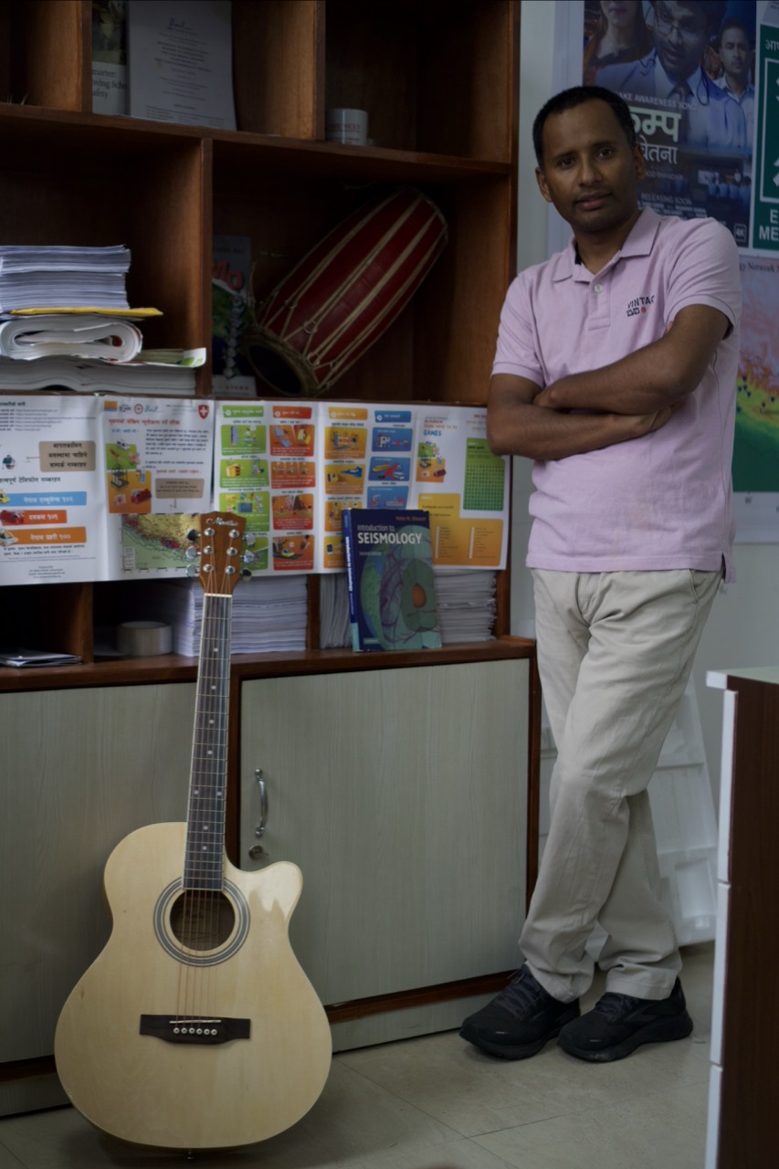 Man in pink polo shirt and beige trousers standing with arms crossed beside acoustic guitar in classroom with bookshelves and educational posters.