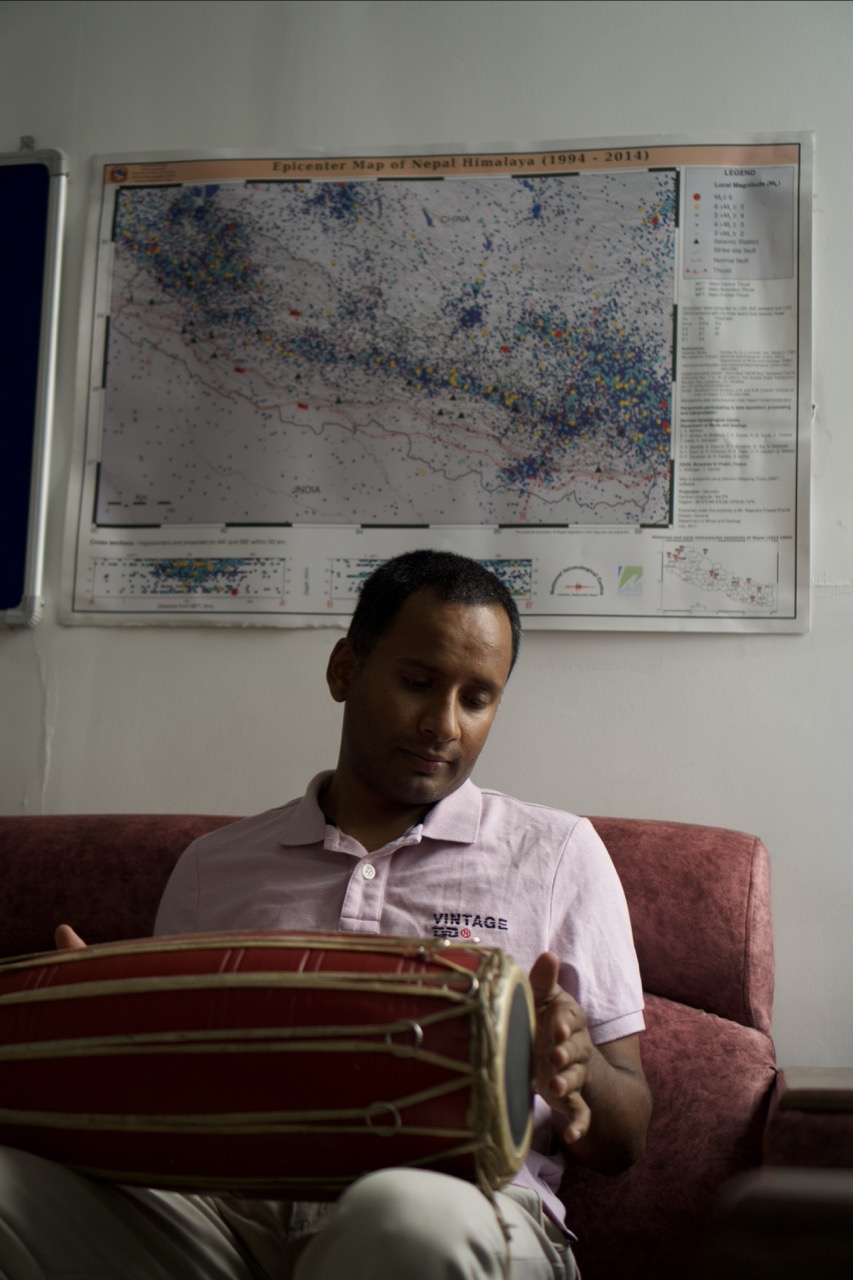 Man in pink polo shirt holding drum whilst seated on red sofa, with large wall map behind him.