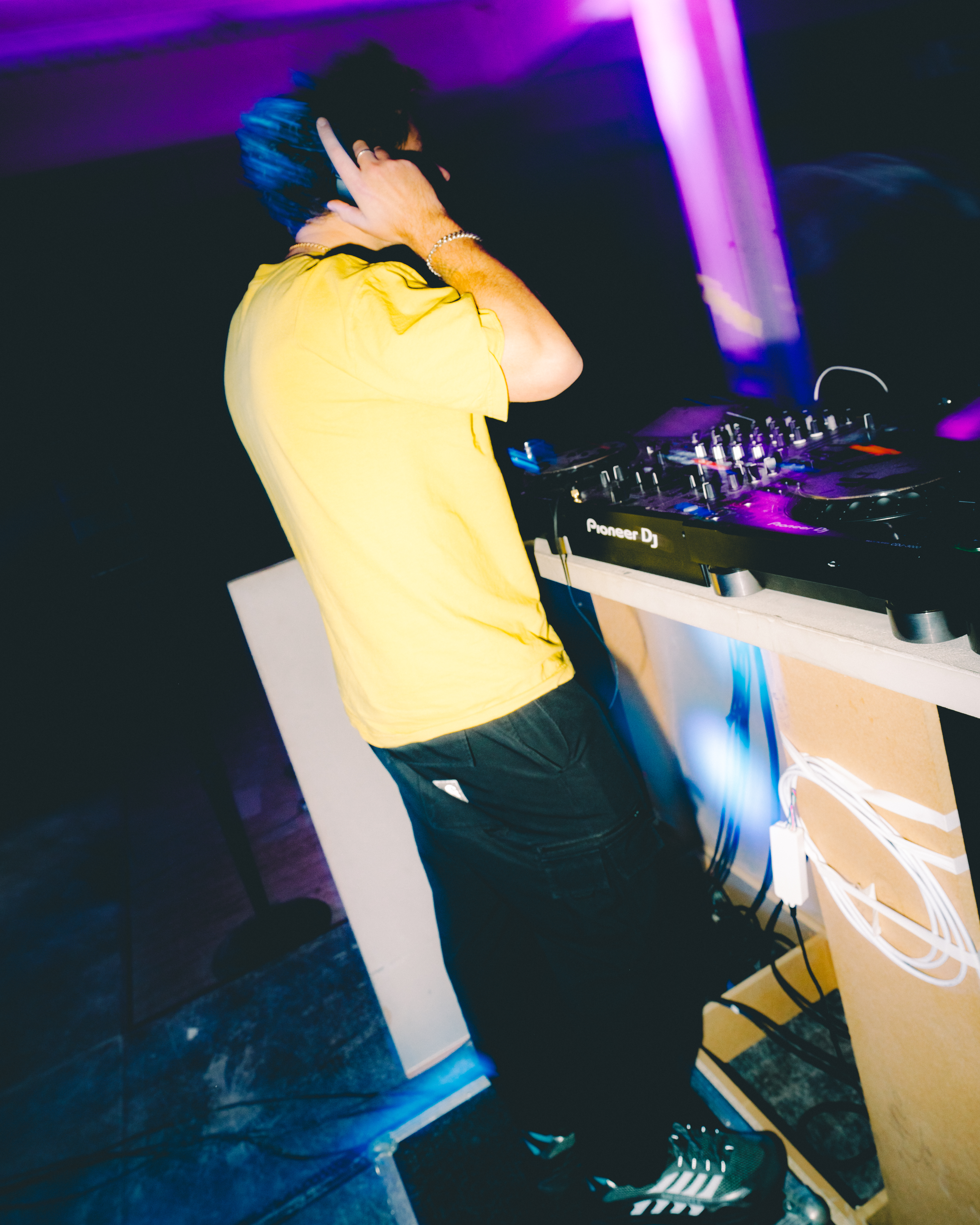 DJ in yellow shirt operating mixing deck with purple and pink neon lighting, one hand on head, standing behind white booth.