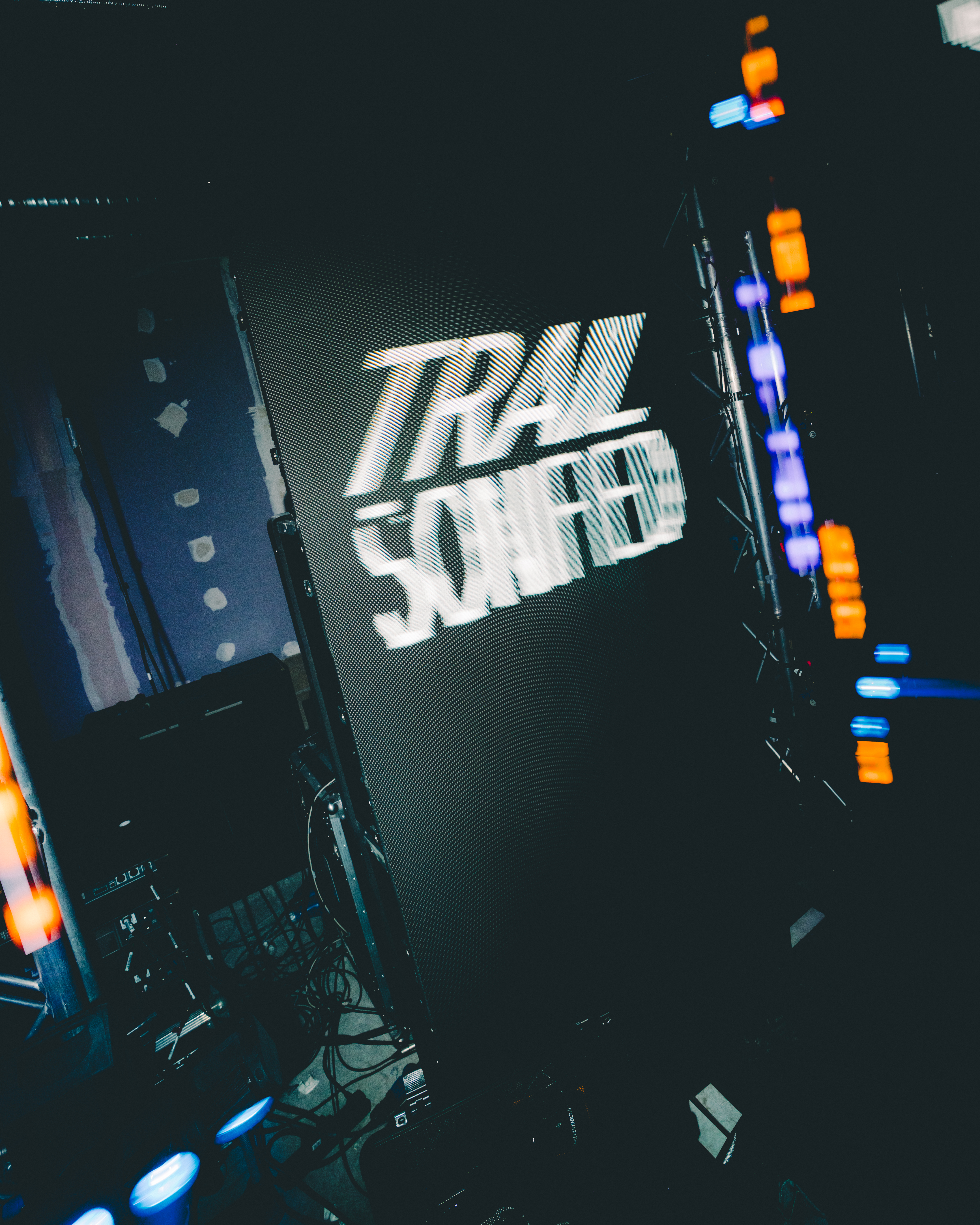 Dark concert stage with "TRAIL SONIC" text in white letters on backdrop, surrounded by blue and orange stage lights and equipment.