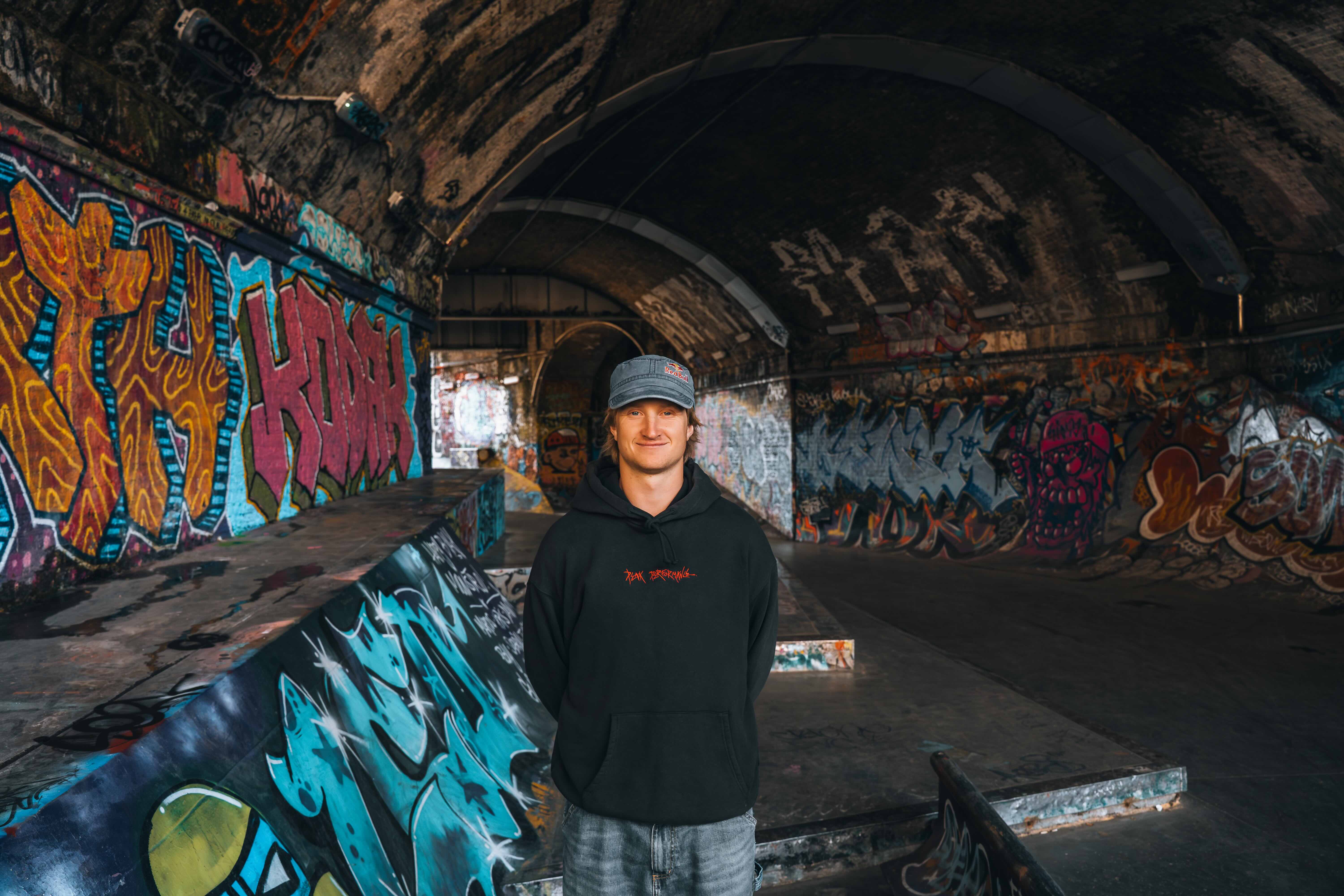 Person in black hoodie and cap standing in graffiti-covered underground tunnel with colourful street art on walls and arched ceiling.
