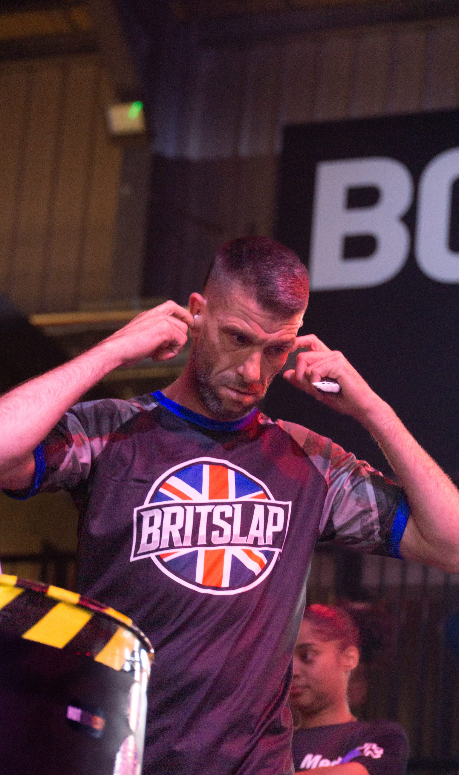 Man in purple camouflage shirt with Union Jack "BRITSLAP" logo adjusting headphones, coloured lighting on stage background.
