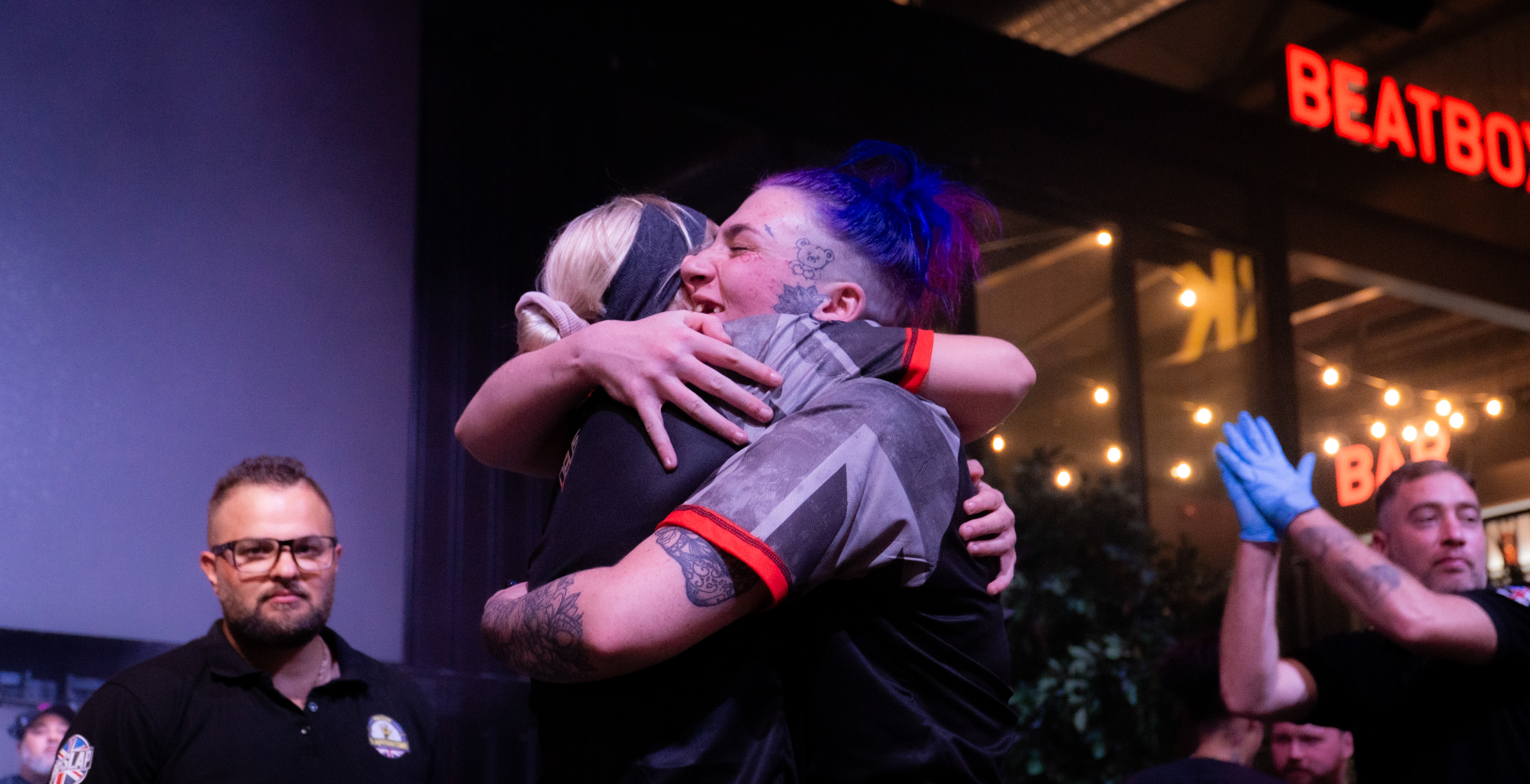 Two men embracing whilst another celebrates with raised hand in background. Red neon "BEATBO" sign visible above dimly lit venue with string lights.