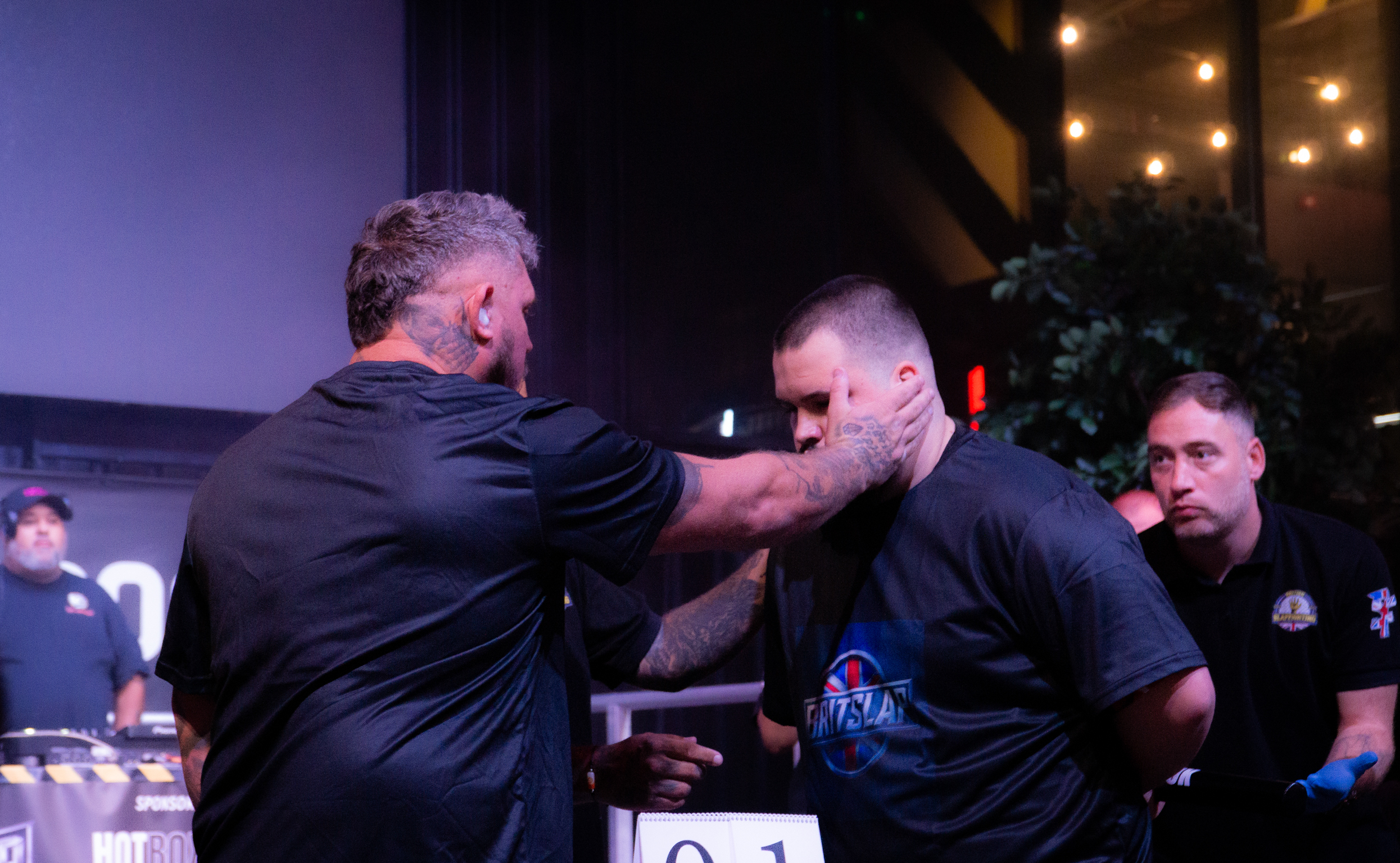 Two men in dark clothing engaged in arm wrestling at a table, with spectators and warm lighting in background.