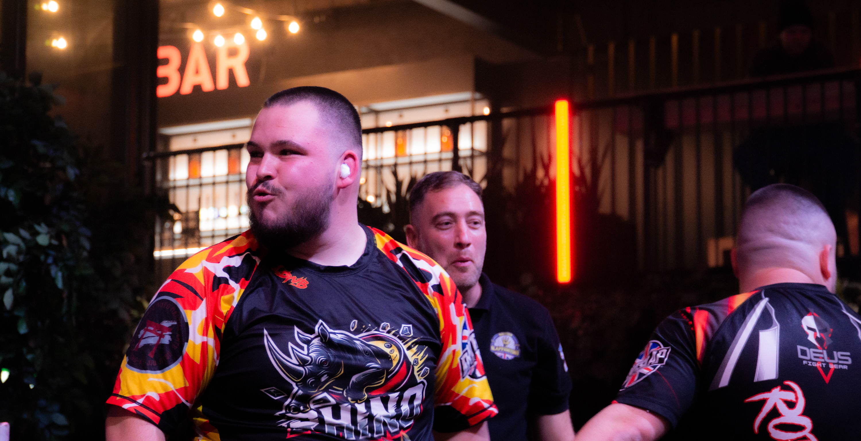 Three men in colourful racing jackets stand outside illuminated bar at night, warm yellow lights visible on building facade.