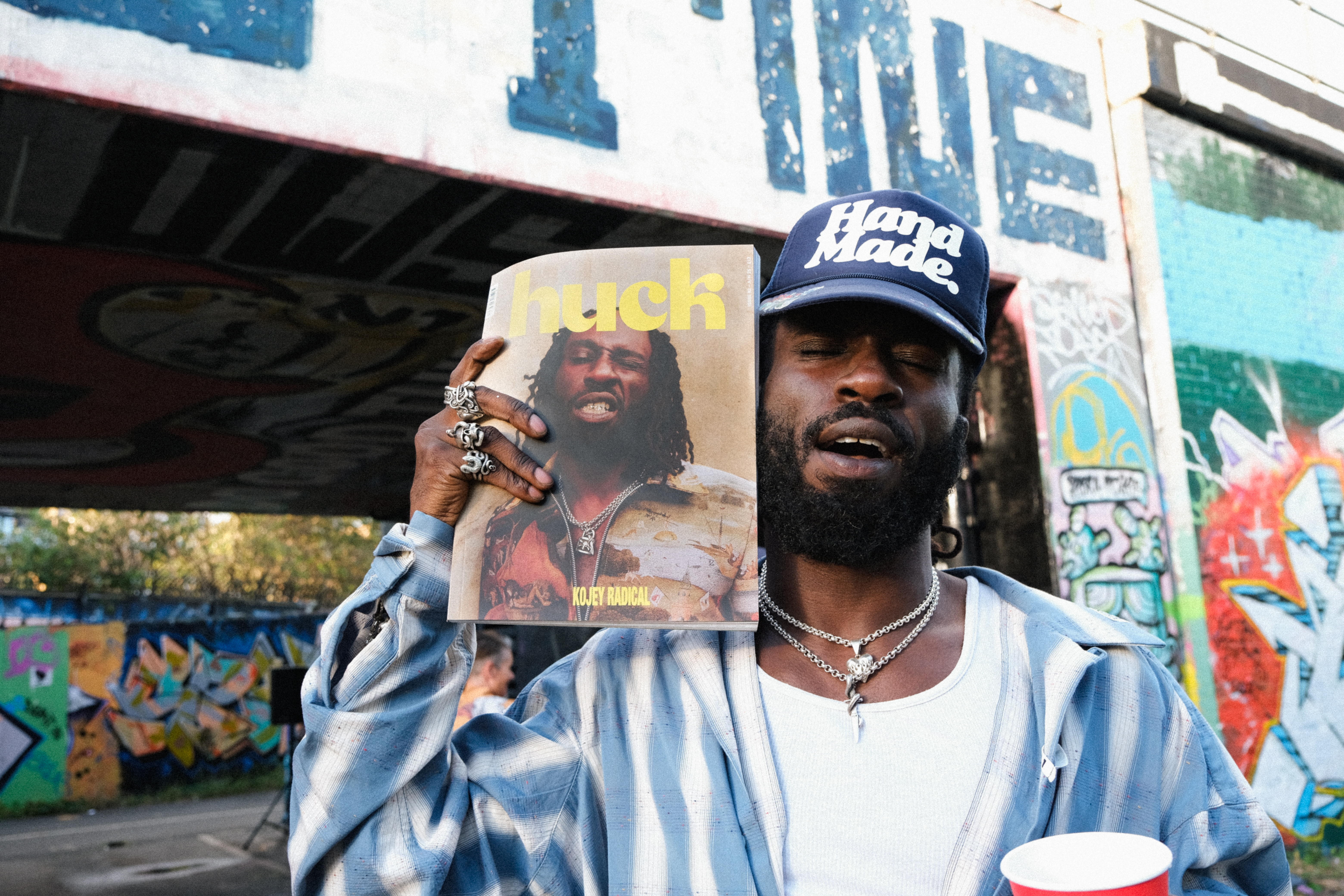Man in blue cap and striped shirt holding magazine, standing against colourful graffitied wall with blue and white painted sections.