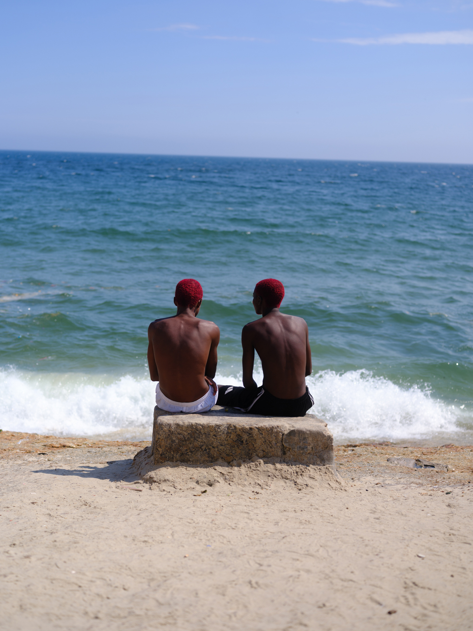 Two shirtless men wearing red caps sit on concrete block at beach, facing turquoise ocean with white waves and blue sky.