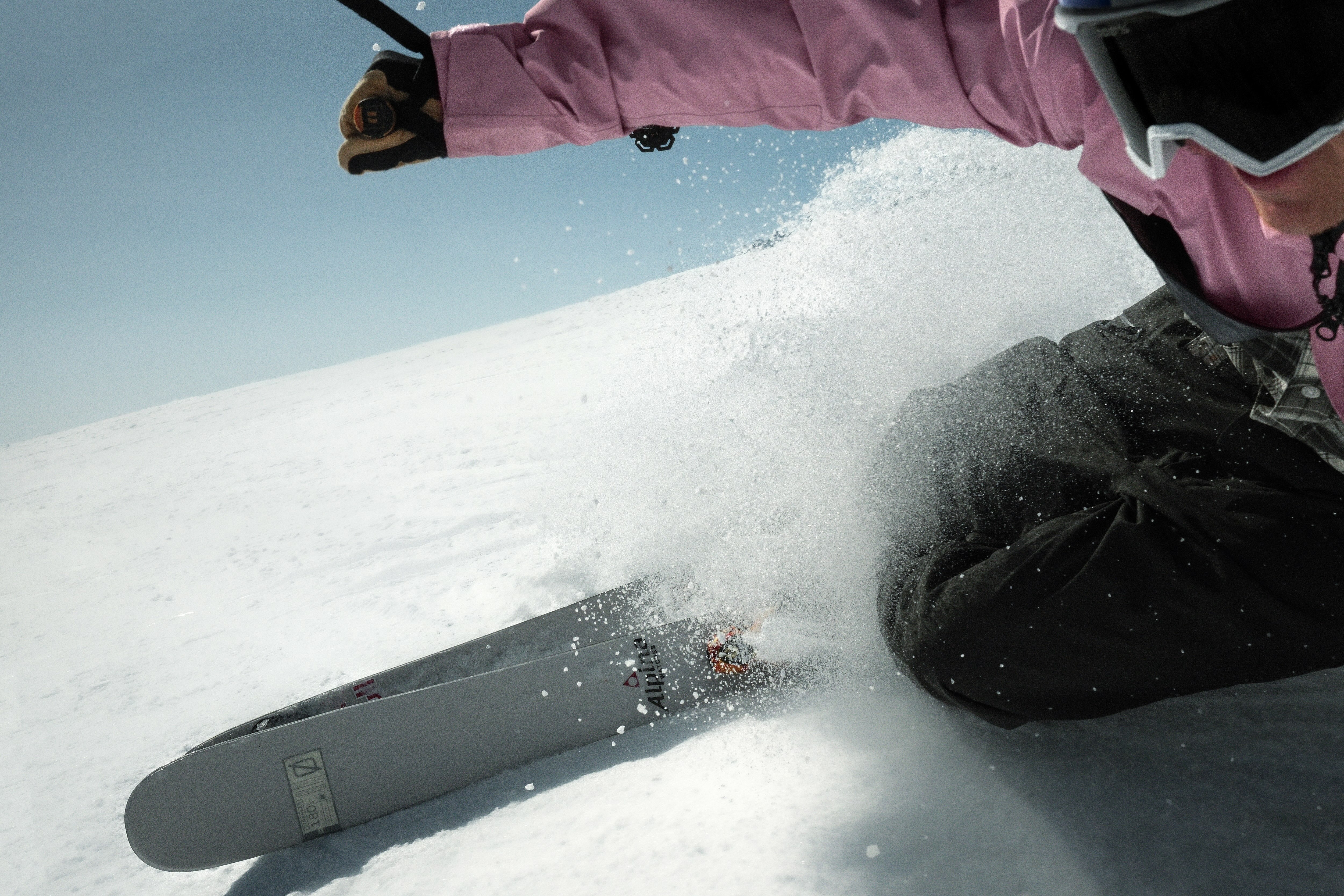 Person in pink jacket kneeling in snow beside grey skis, powder snow spraying up against blue sky background.