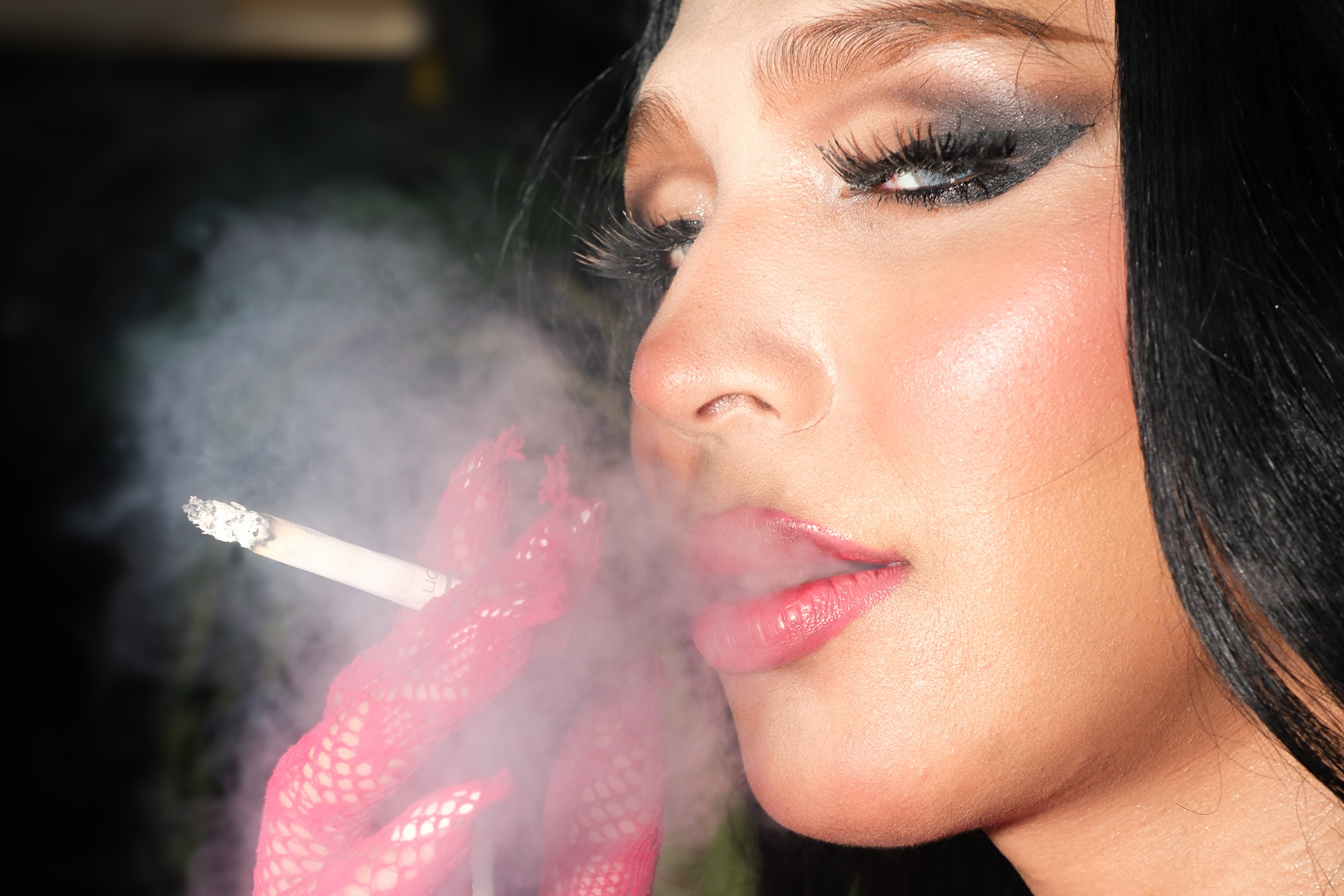 Woman with dark hair and pink lips exhaling white smoke, eyes closed, face illuminated against dark background.