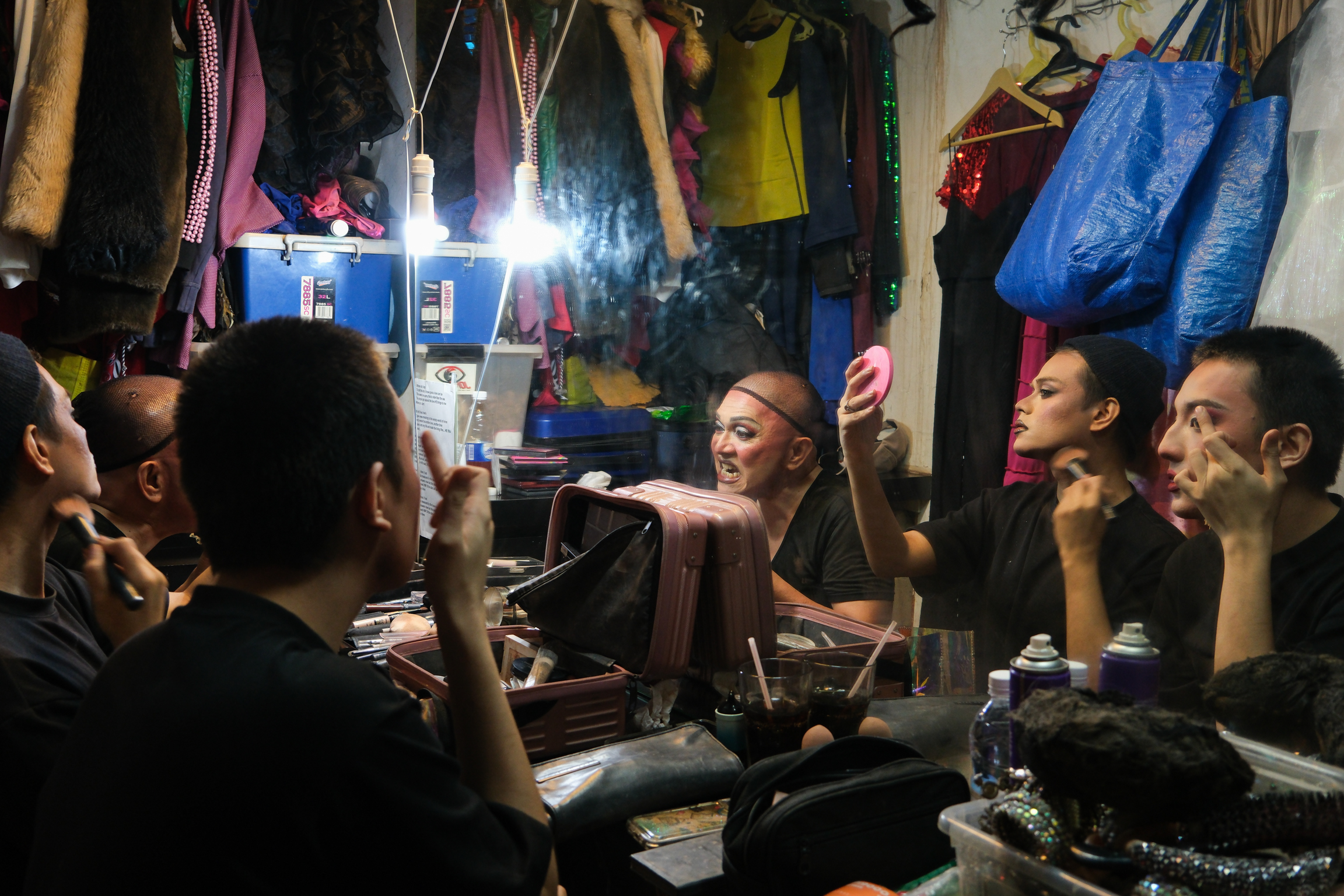Group of men sitting in cramped indoor space with hanging clothes, bags, and belongings. Bright light illuminates centre of cluttered room.