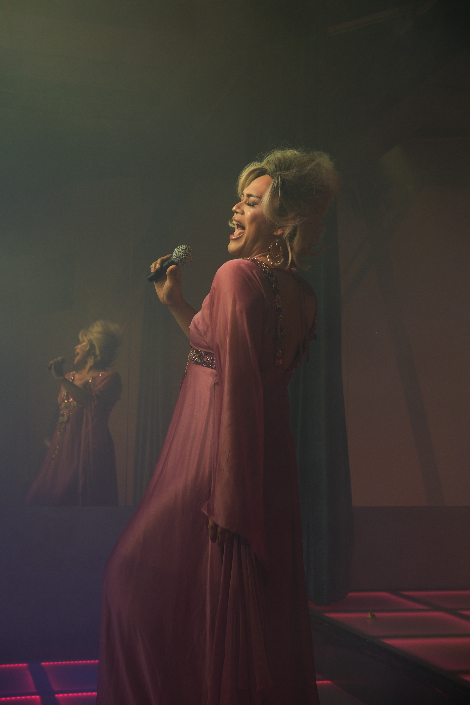 Woman in burgundy dress singing into microphone on dimly lit stage with atmospheric lighting and shadowy figure in background.
