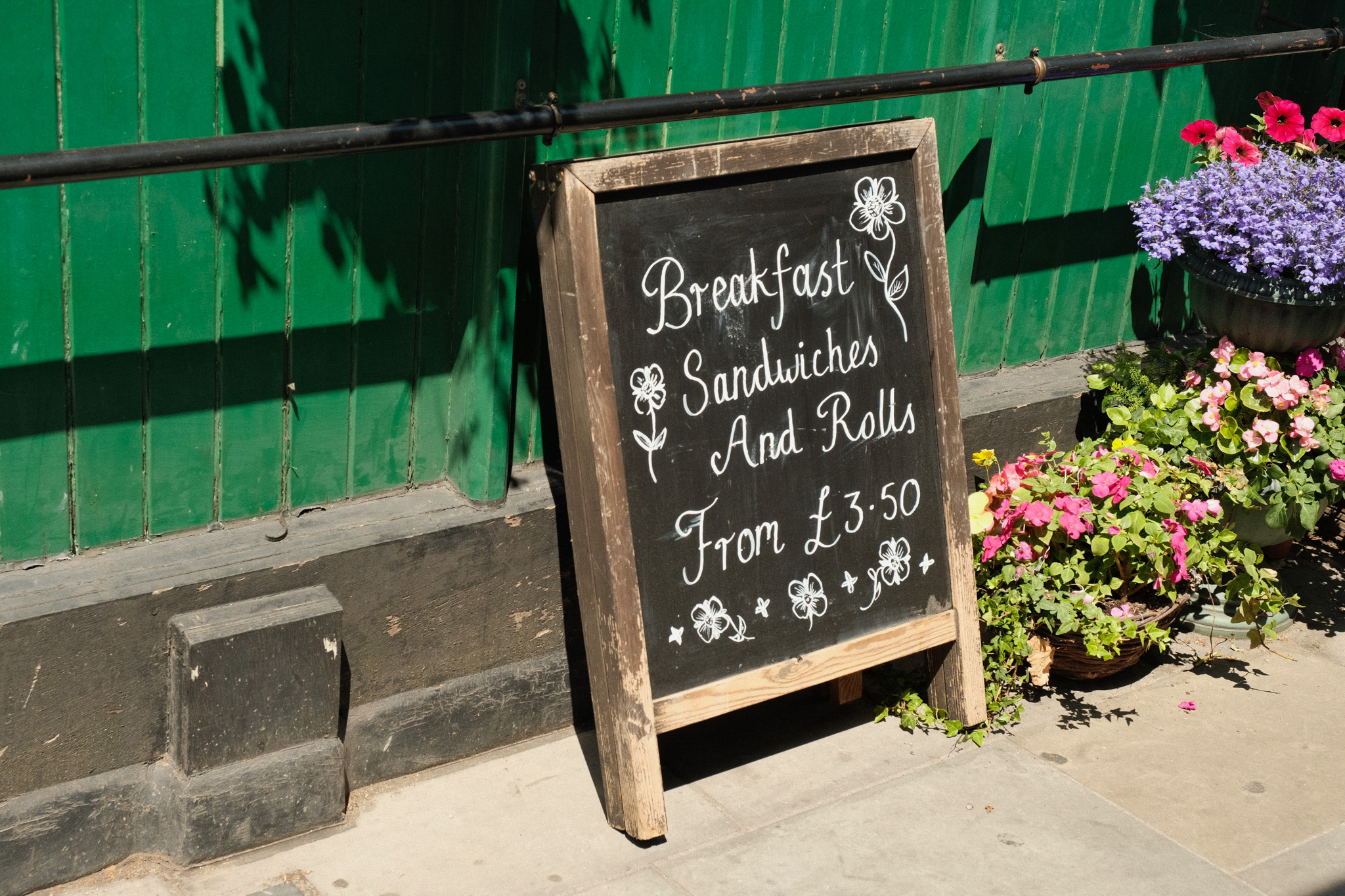 Black chalkboard sign with white text advertising breakfast sandwiches and rolls from £3.50, propped against green wooden building with colourful flowers.