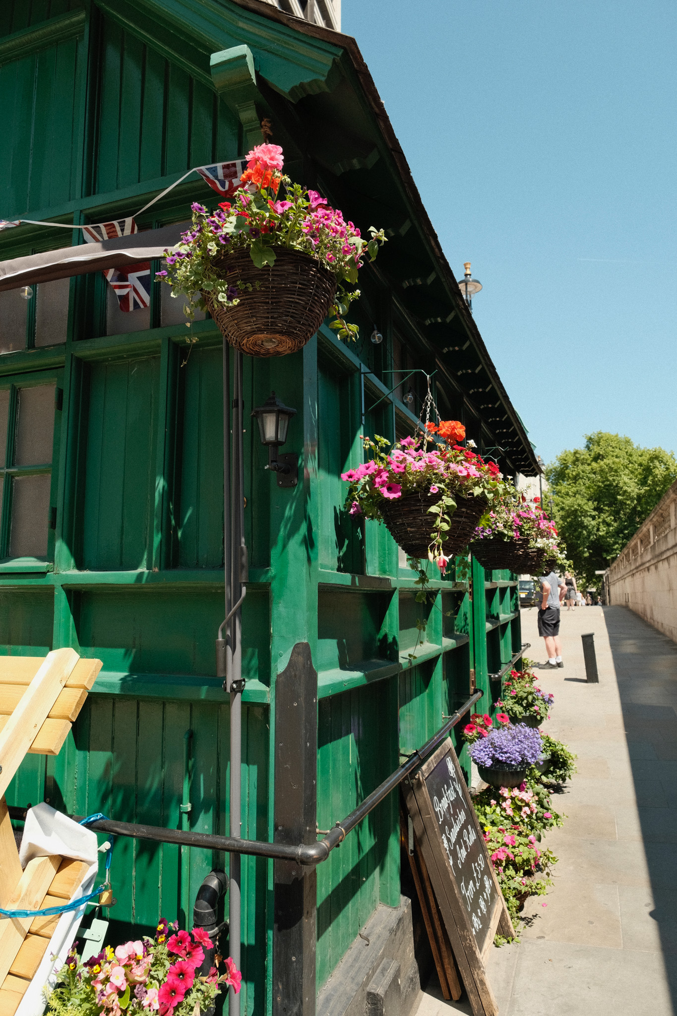 Green timber-framed building with black balconies decorated with hanging baskets of pink and purple flowers against blue sky.