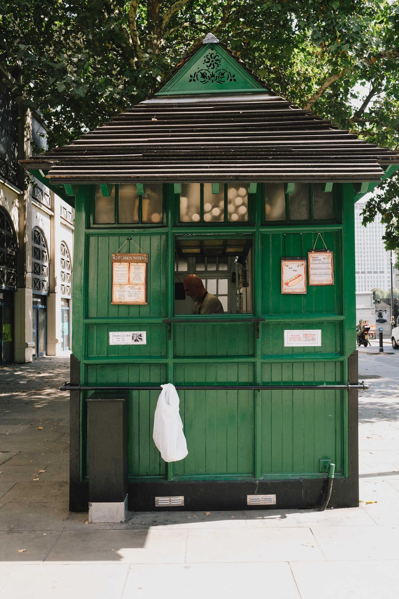 Green wooden Victorian kiosk with triangular roof and multiple windows displaying menus, located on tree-lined pavement.