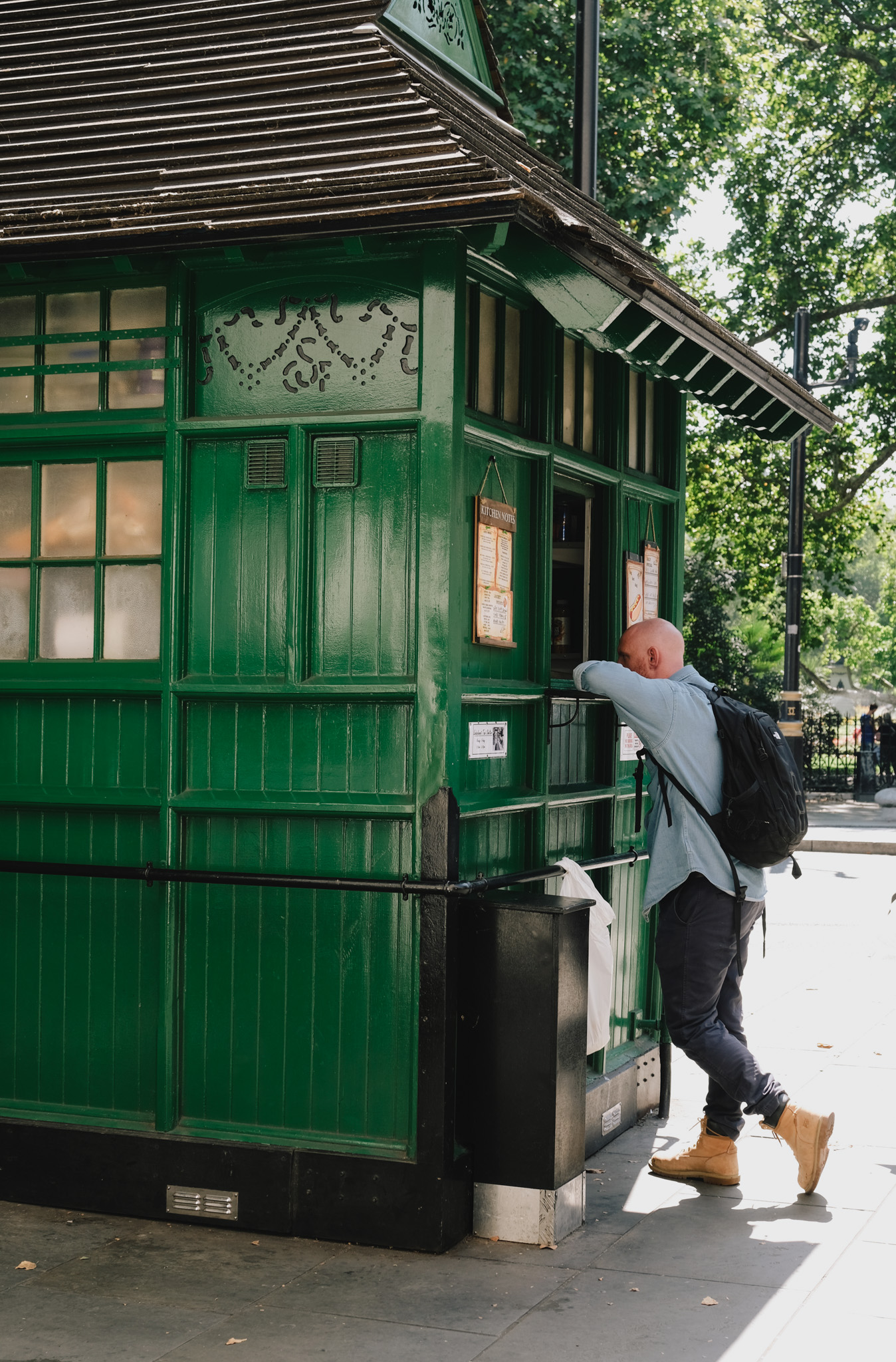 Person in grey hoodie and rucksack looking into green wooden Victorian-style kiosk with decorative trim, surrounded by trees on pavement.