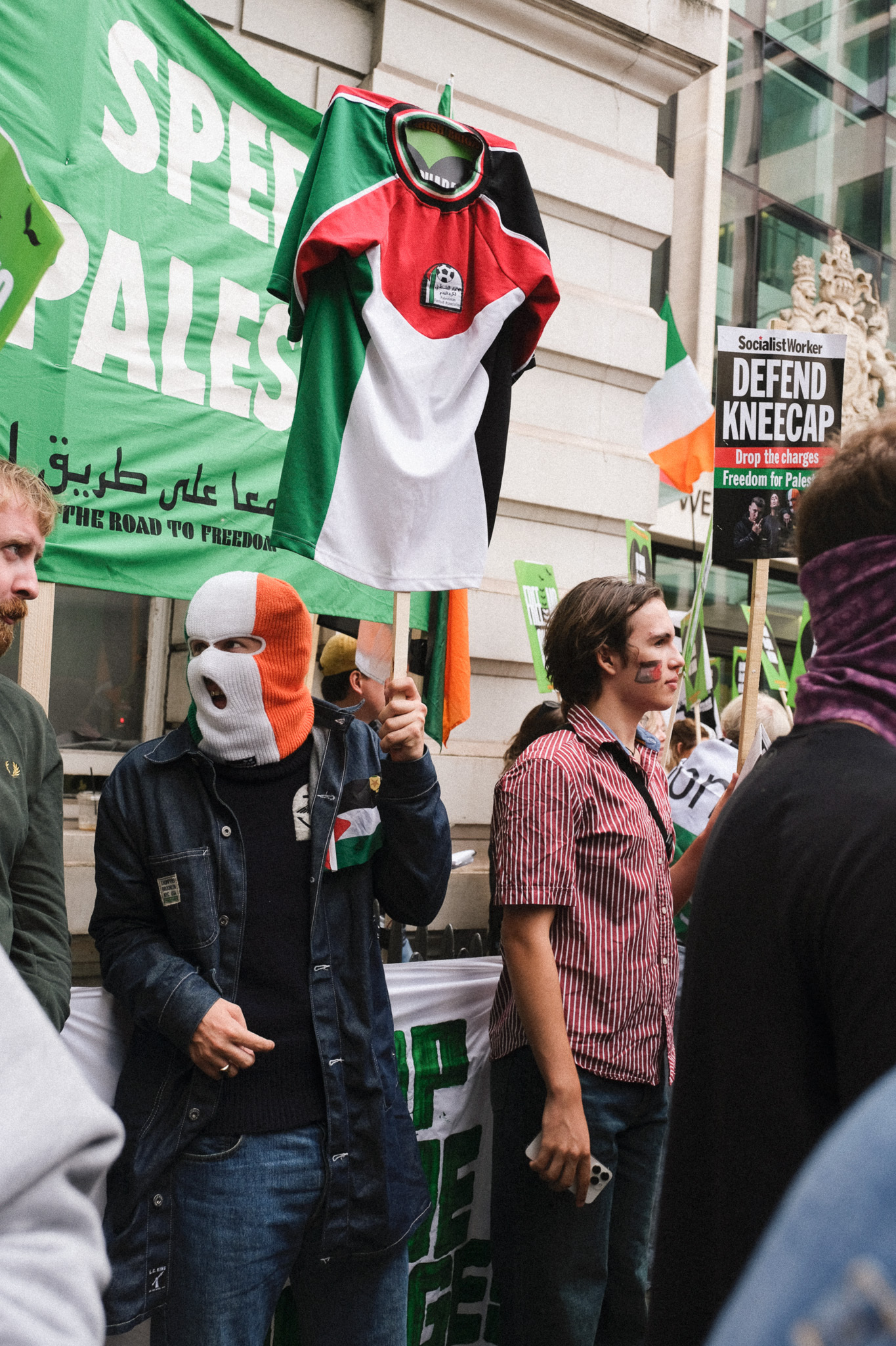 Protesters holding Palestinian flags and signs, one person wearing Irish tricolour face mask, man in striped shirt amongst crowd.