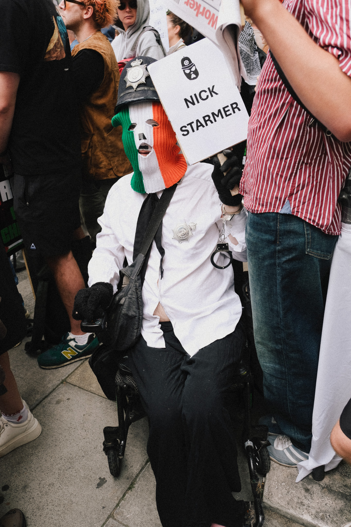 Person in white shirt and mask holding "NICK STARMER" sign at crowded outdoor gathering, surrounded by people in casual clothing.