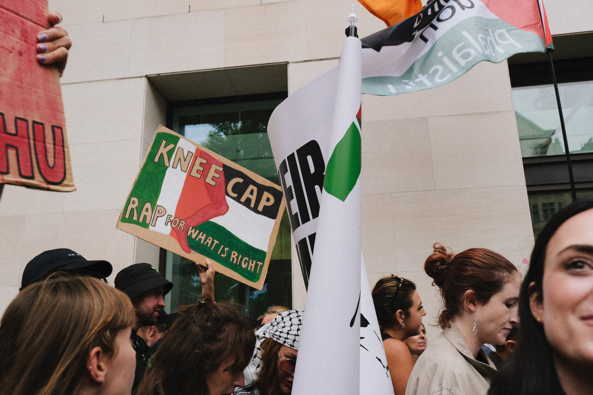 Crowd holding protest signs including "KNEE CAP" and Palestinian flags outside building with glass windows.