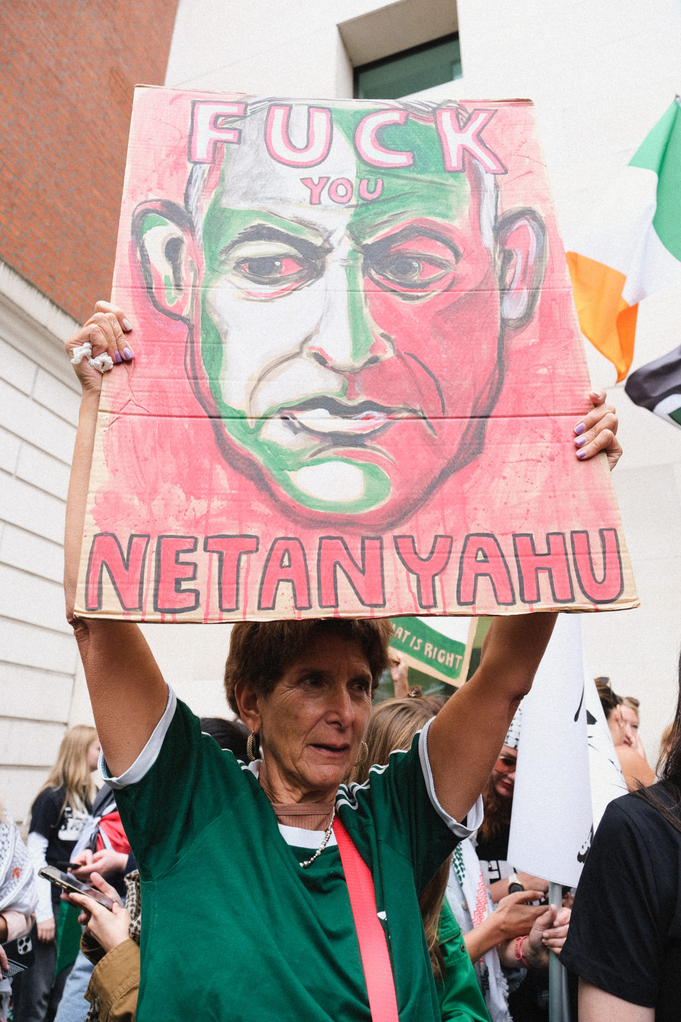 Woman in green shirt holding protest sign with painted portrait and text "FUCK YOU NETANYAHU" in pink and green colours.