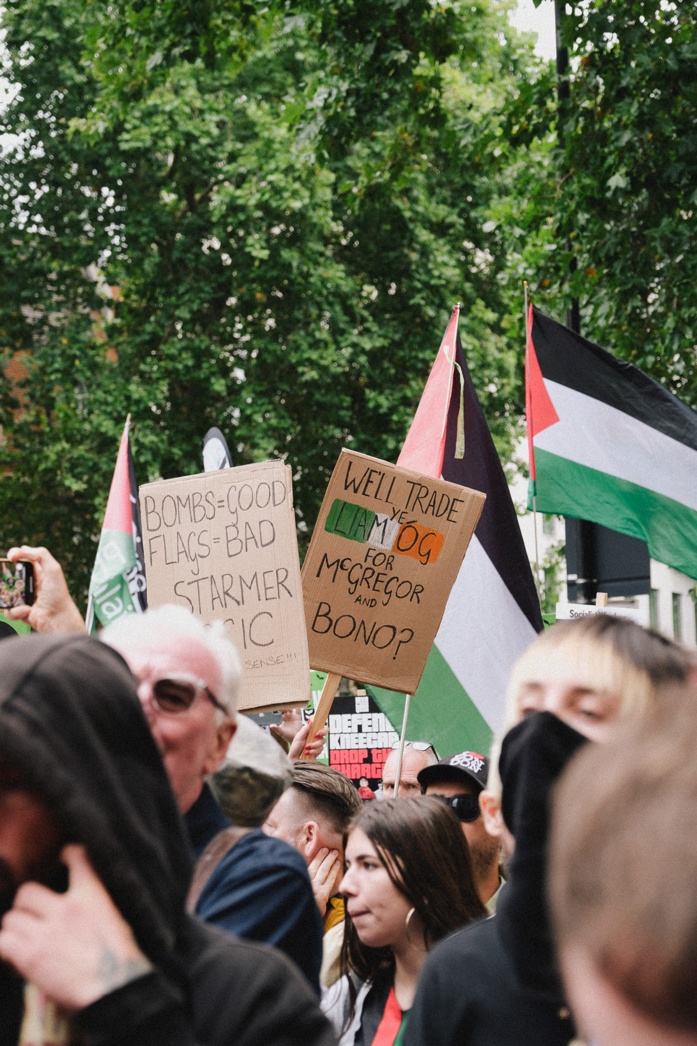 Crowd holding Palestinian flags and protest signs including "BOMBS=GOOD FLATS=BAD STARMER" and "WELL TRADE KEIR OR McGREGOR BONOP" beneath green trees.