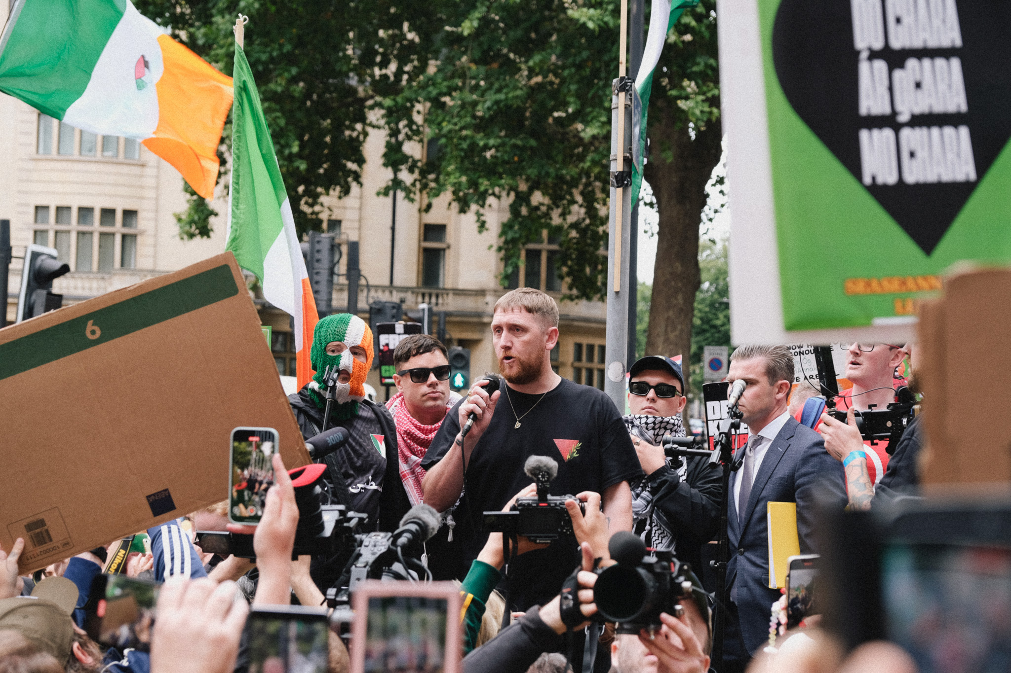 Man in black shirt speaking at microphones surrounded by crowd holding Irish flags and banners on street.
