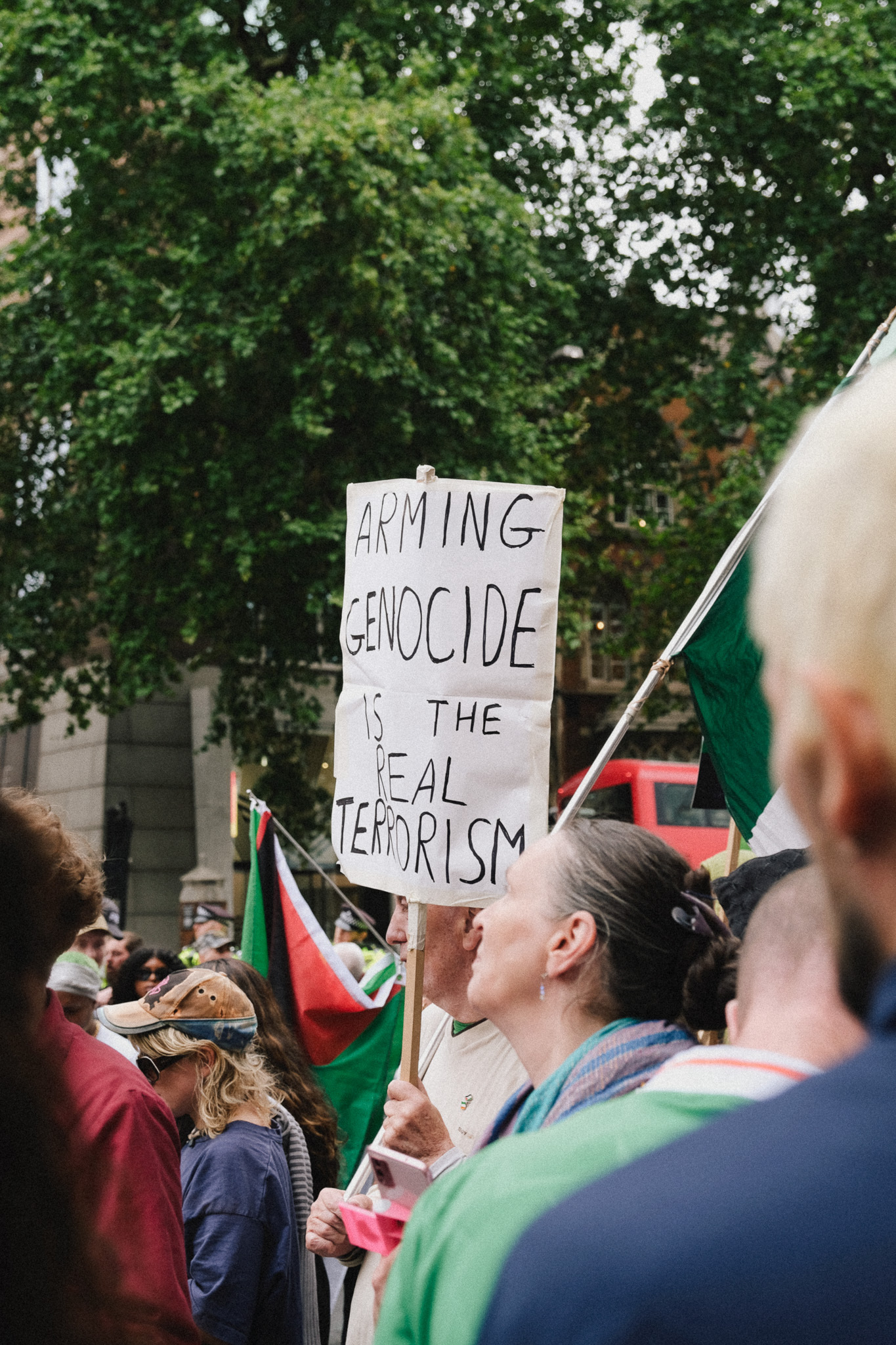 Protest crowd with person holding white placard reading "ARMING GENOCIDE IS THE REAL TERRORISM" amongst Palestinian flags and green foliage.