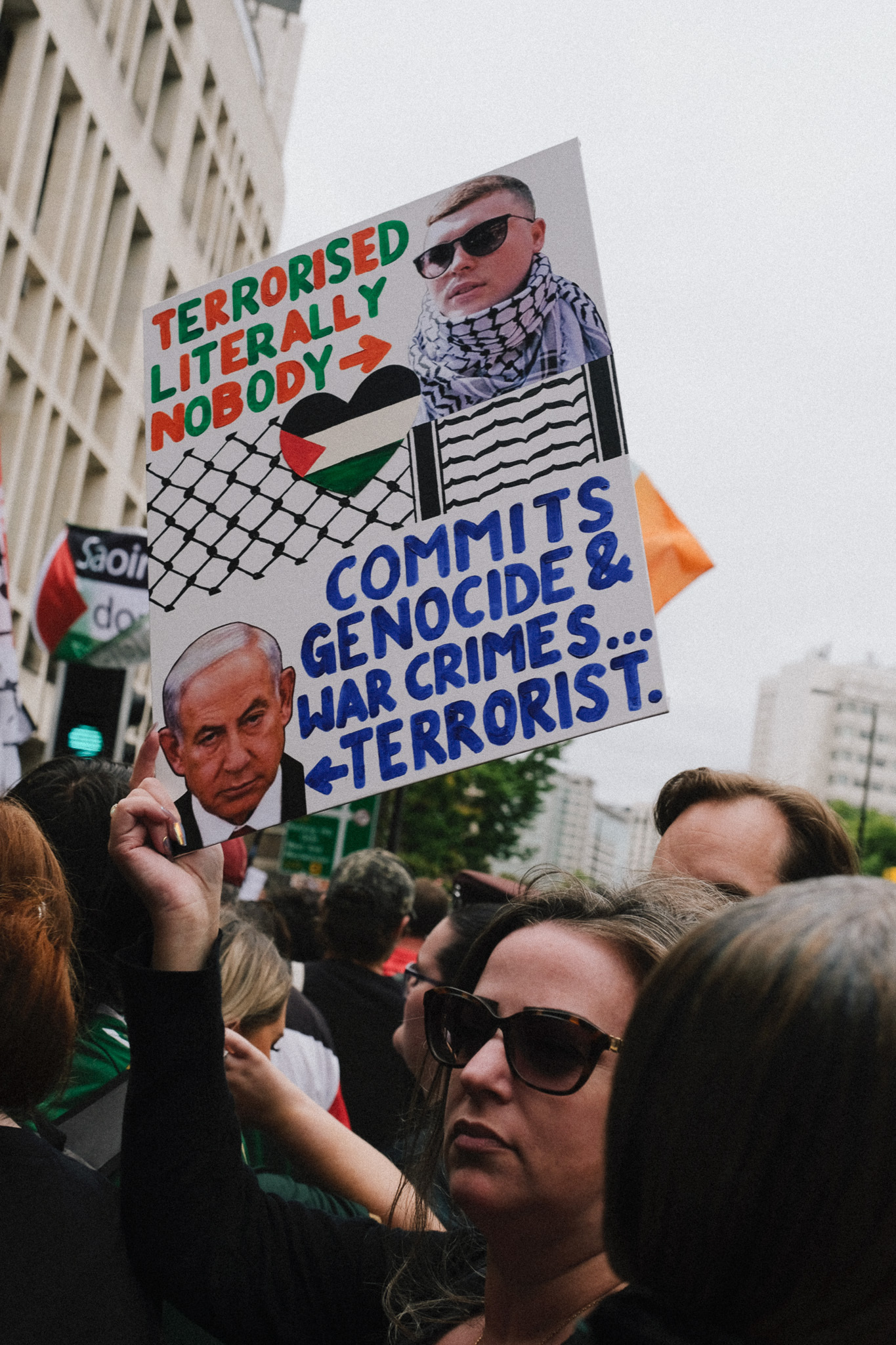 Protest sign with colourful text "TERRORISED LITERALLY NOBODY" and blue text "COMMITS GENOCIDE & WAR CRIMES... TERRORIST" held above crowd.