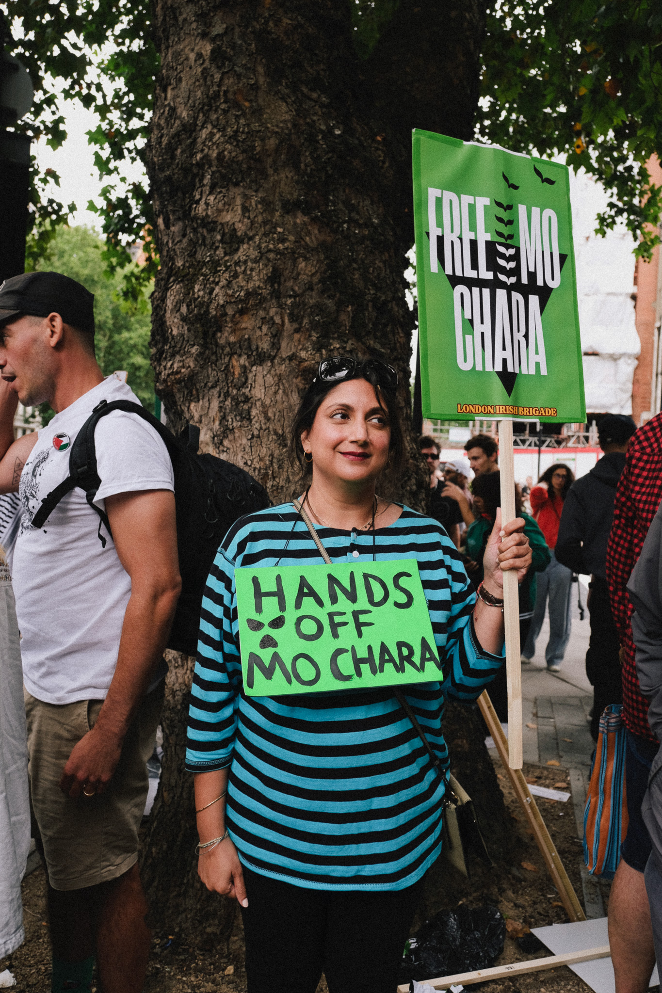 Woman in blue striped top holds green "HANDS OFF MO CHARA" sign at outdoor protest, with "FREE MO CHARA" banner visible behind.