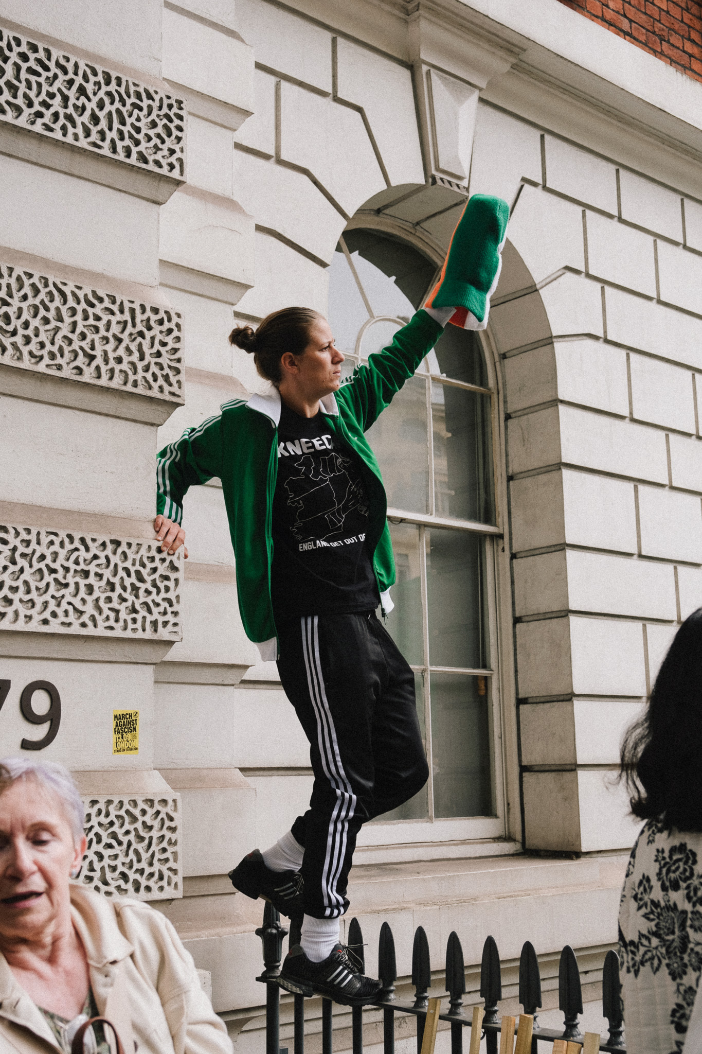 Woman in green tracksuit jumping with arm raised outside white Georgian building with decorative stonework and iron railings.