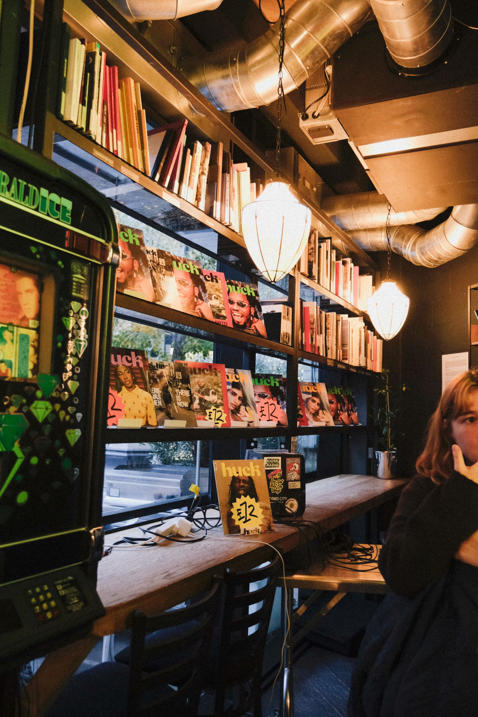 Interior of bookshop with shelves of books, magazines in window display, exposed metallic ductwork on ceiling, warm lighting, and person browsing.