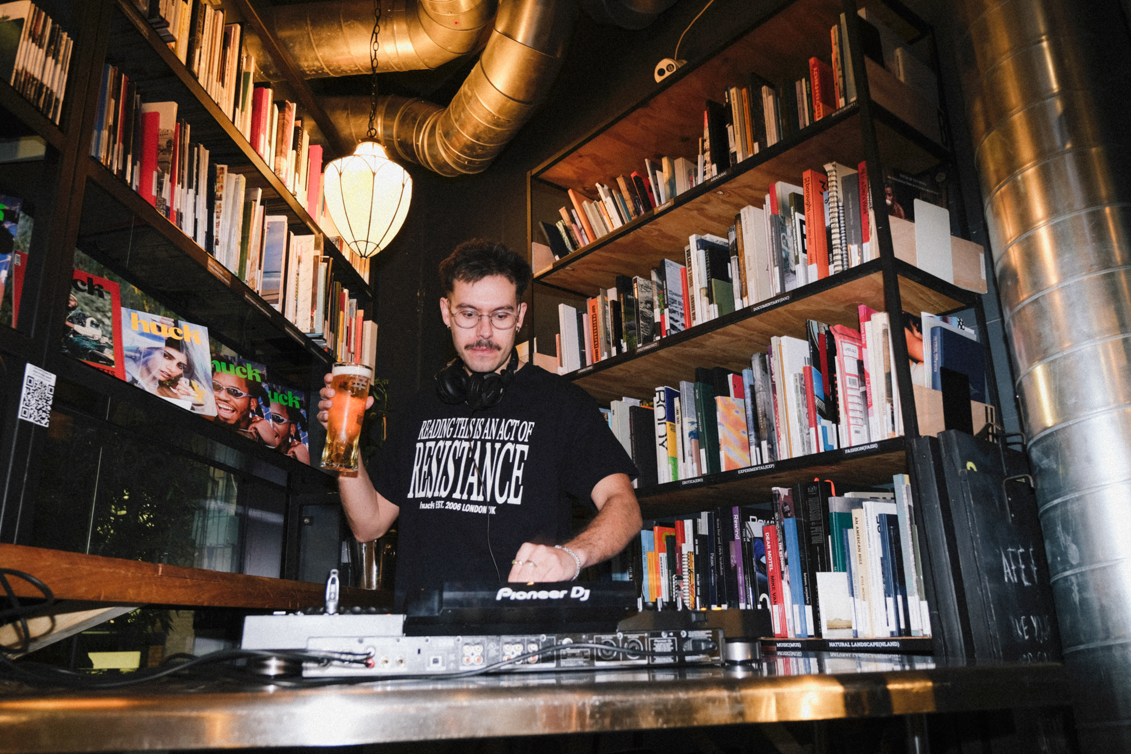 DJ in black t-shirt mixing music at turntables, surrounded by floor-to-ceiling bookshelves, holding pint glass, exposed ductwork overhead.