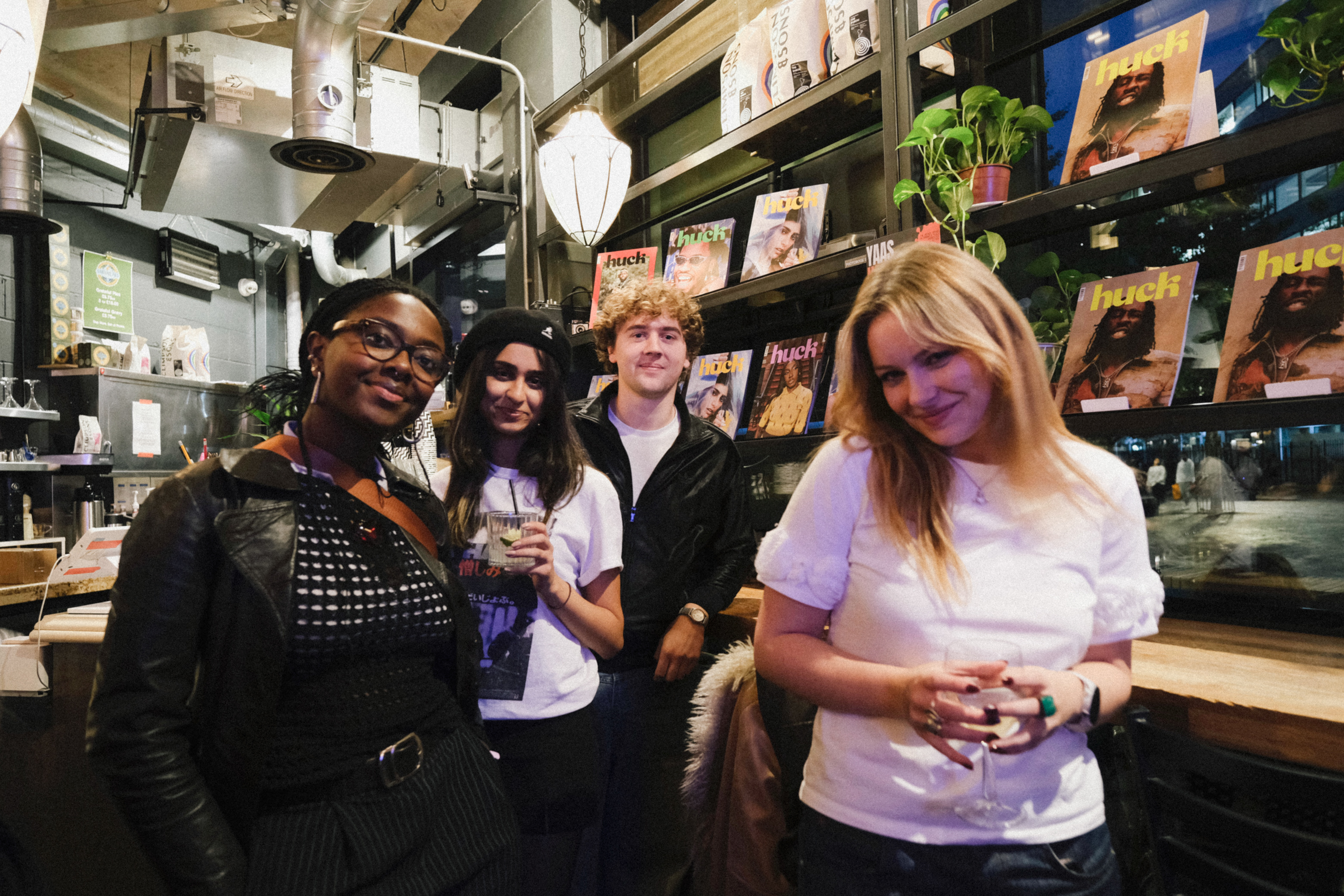 Four people standing in record shop with vinyl albums on shelves behind them, holding drinks and smiling at camera.