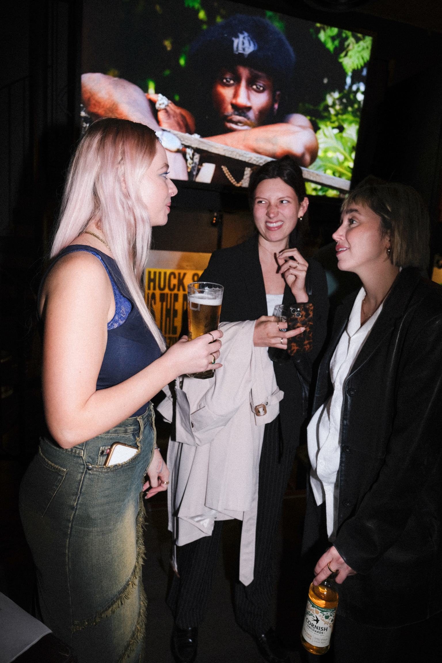 Three women socialising with drinks in dimly lit venue beneath large screen displaying film scene with man holding weapon.