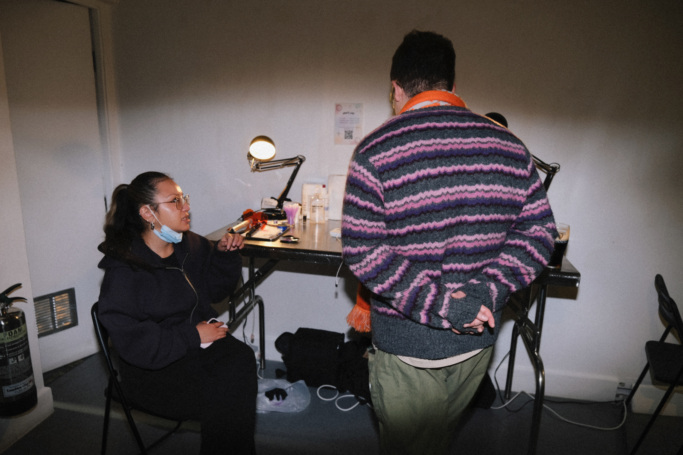 Woman wearing face mask at desk with lamp facing man in purple striped jumper, both seated at black table in white room.