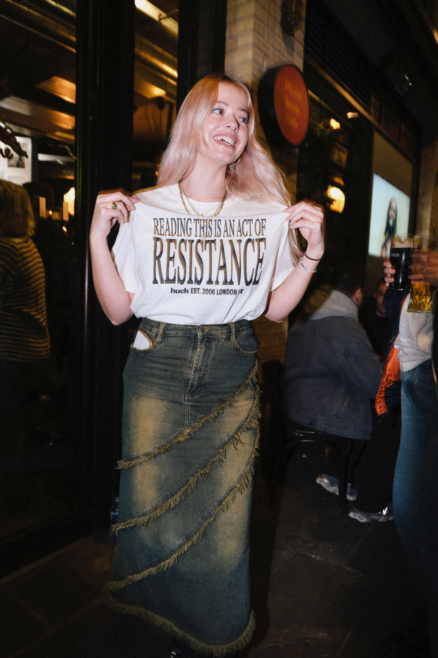 Woman with long blonde hair holding white t-shirt reading "Reading this is an act of resistance" in restaurant interior.