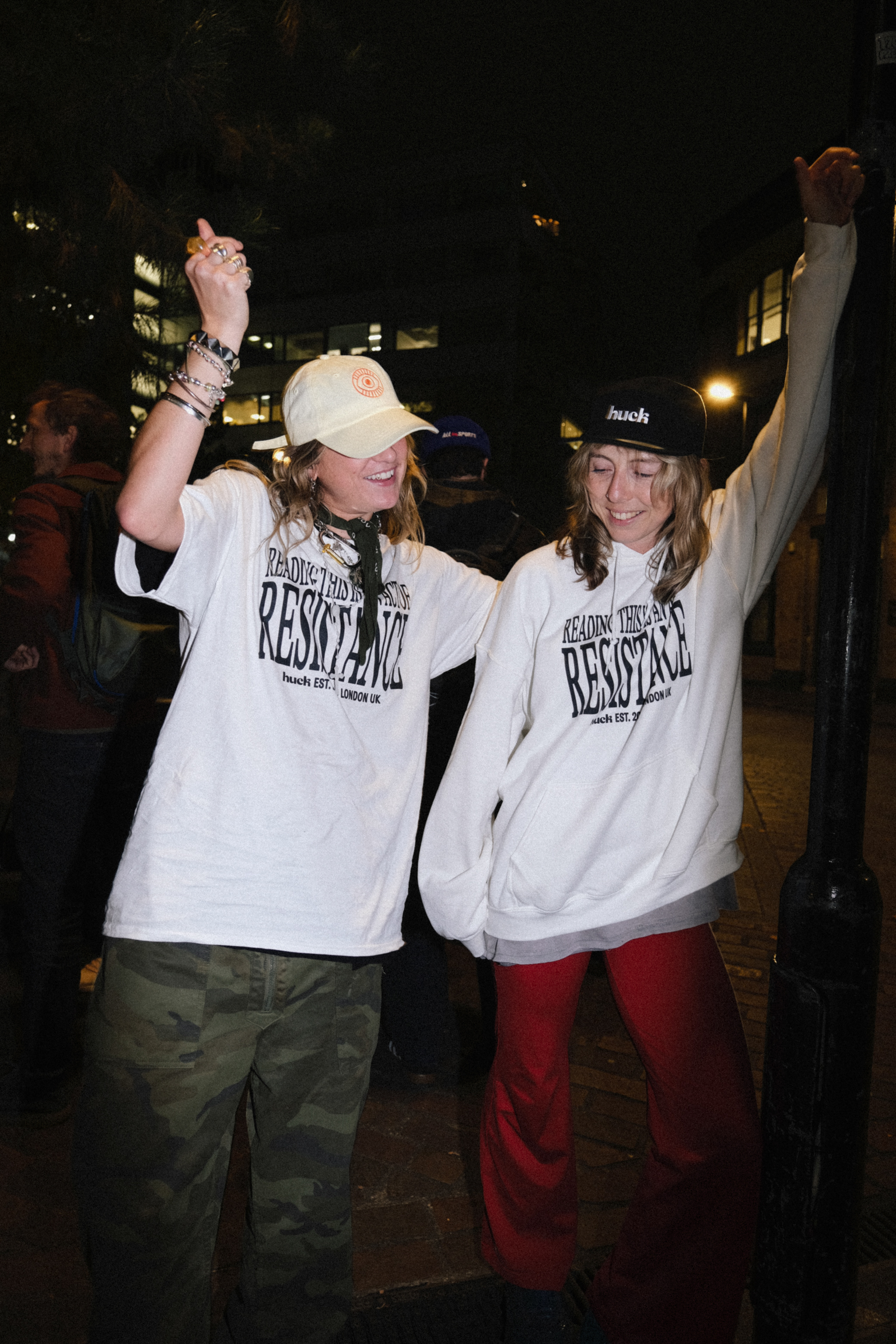 Two people celebrating with raised arms, wearing white "RESOLVED" t-shirts and caps, standing on a street at night with buildings behind.