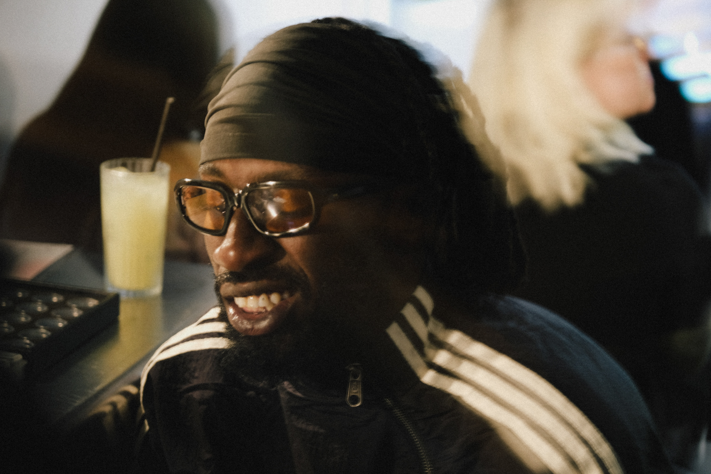 Black man wearing glasses and beanie smiling, wearing dark jacket with white stripes, yellow drink with straw nearby, blurred figures behind