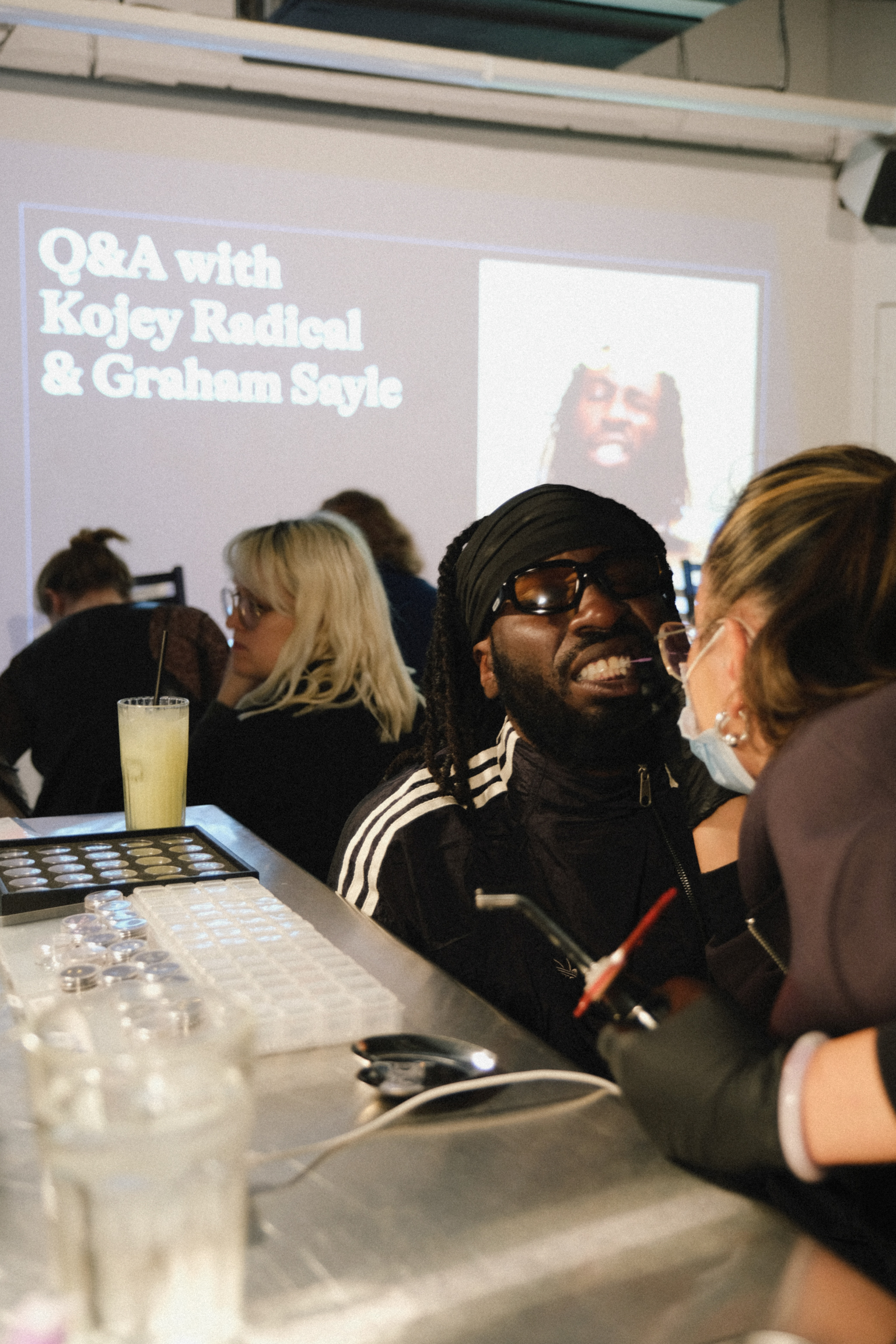 Man with dreadlocks and sunglasses smiling at Q&A event, seated at table with others, projection screen shows "Q&A with Kojey Radical & Graham Sayle"