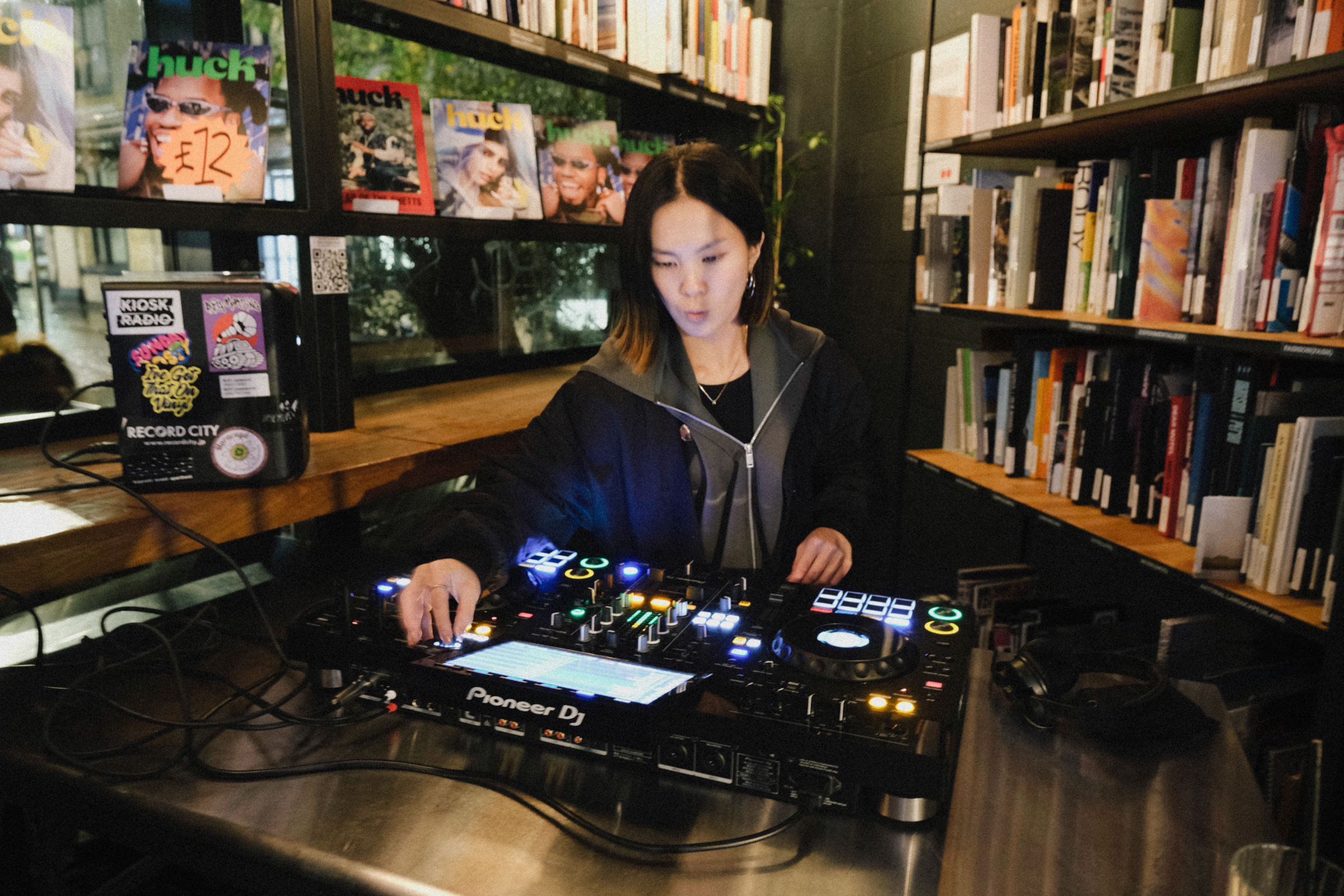 Woman using DJ mixing console with blue LED lights in bookshop surrounded by bookshelves and magazines.