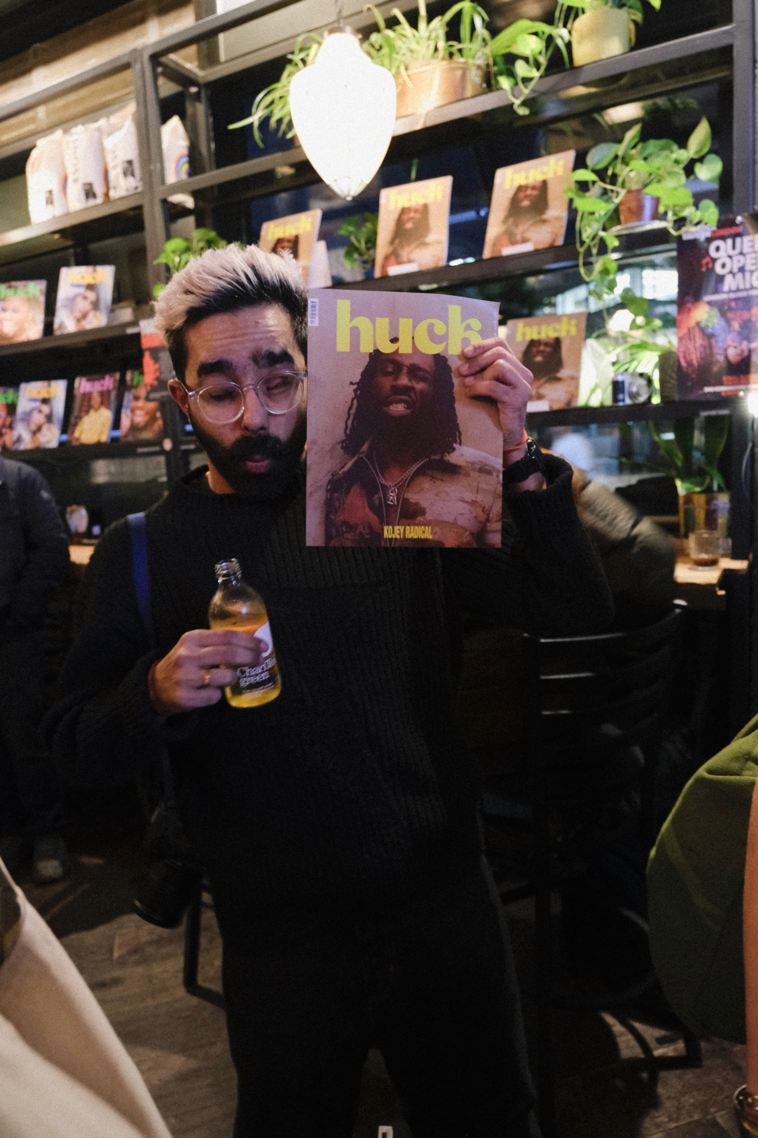 Bearded man in glasses holds Huck magazine and beer bottle in bookshop with shelves of books and hanging plants behind him.