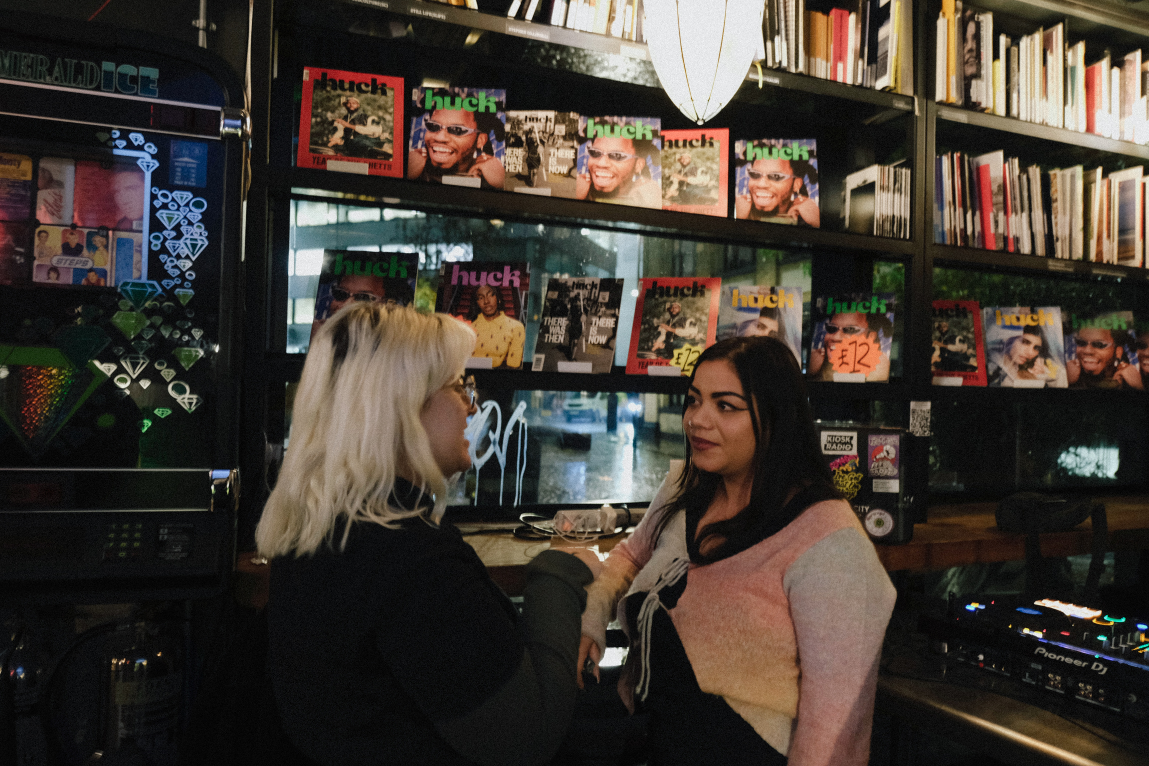 Two women at newsstand counter with magazines displayed on shelves behind them, books visible on upper right shelving.