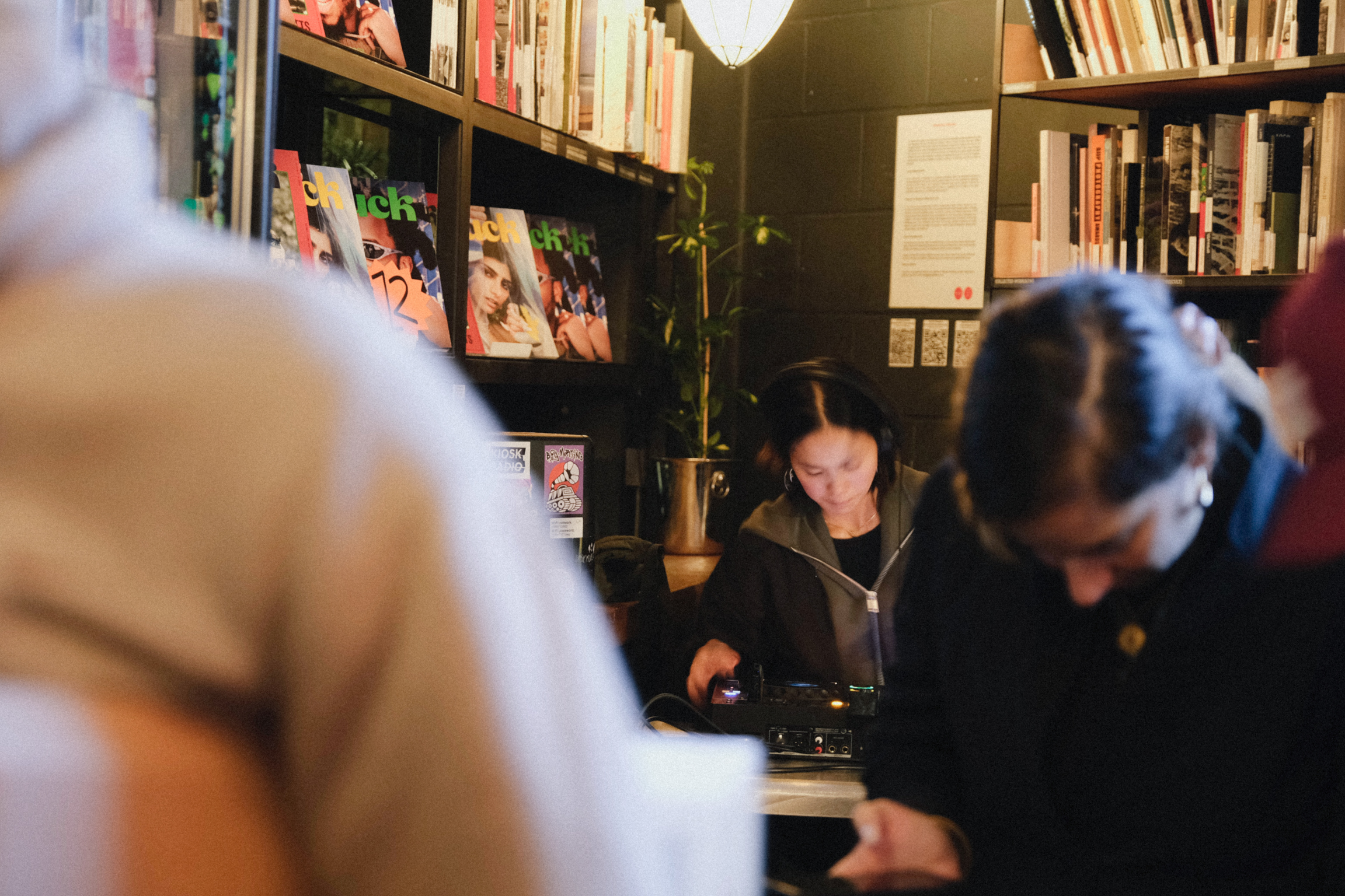 Woman sitting at table in bookshop surrounded by bookshelves, with blurred figures in foreground.
