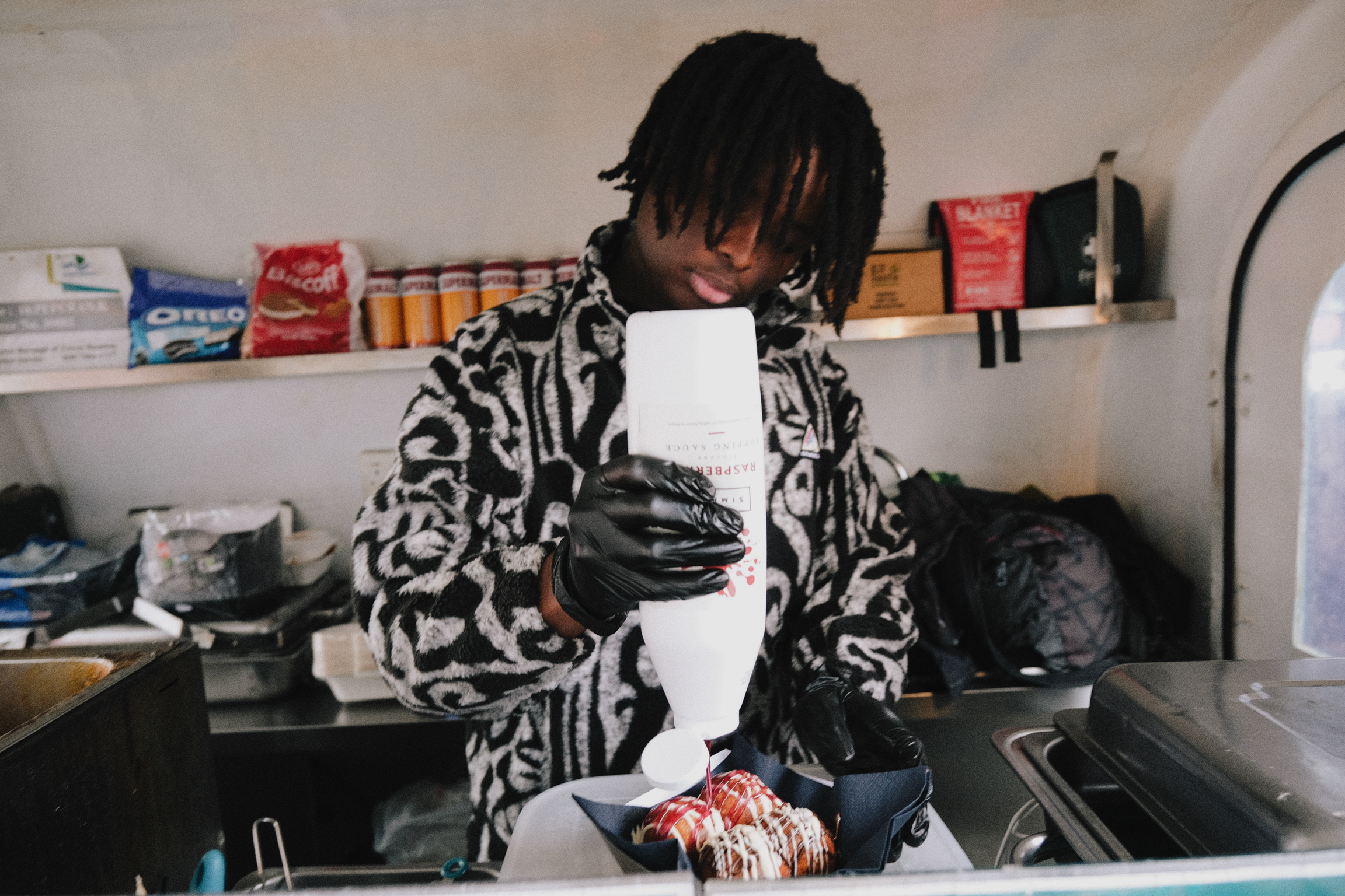 Person with dreadlocks wearing black and white patterned hoodie cooking in kitchen, holding white paper, shelves with food items visible.