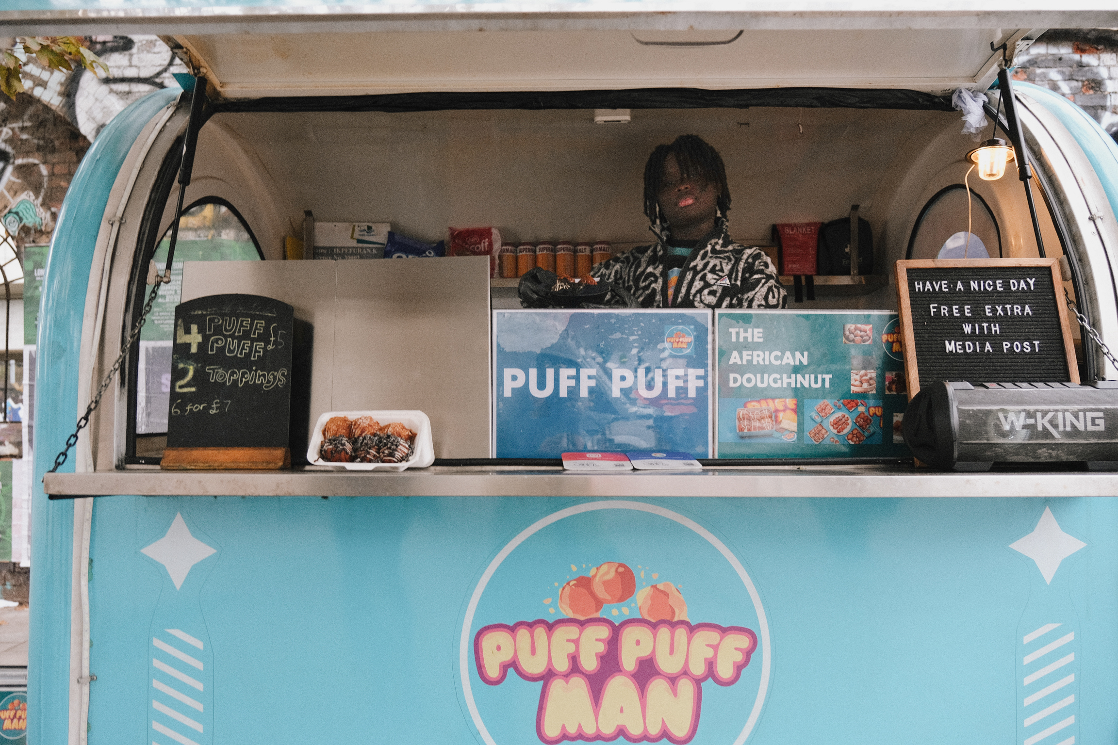 Man standing in turquoise food van with "PUFF PUFF MAN" branding, serving window open, menu boards and equipment visible inside.