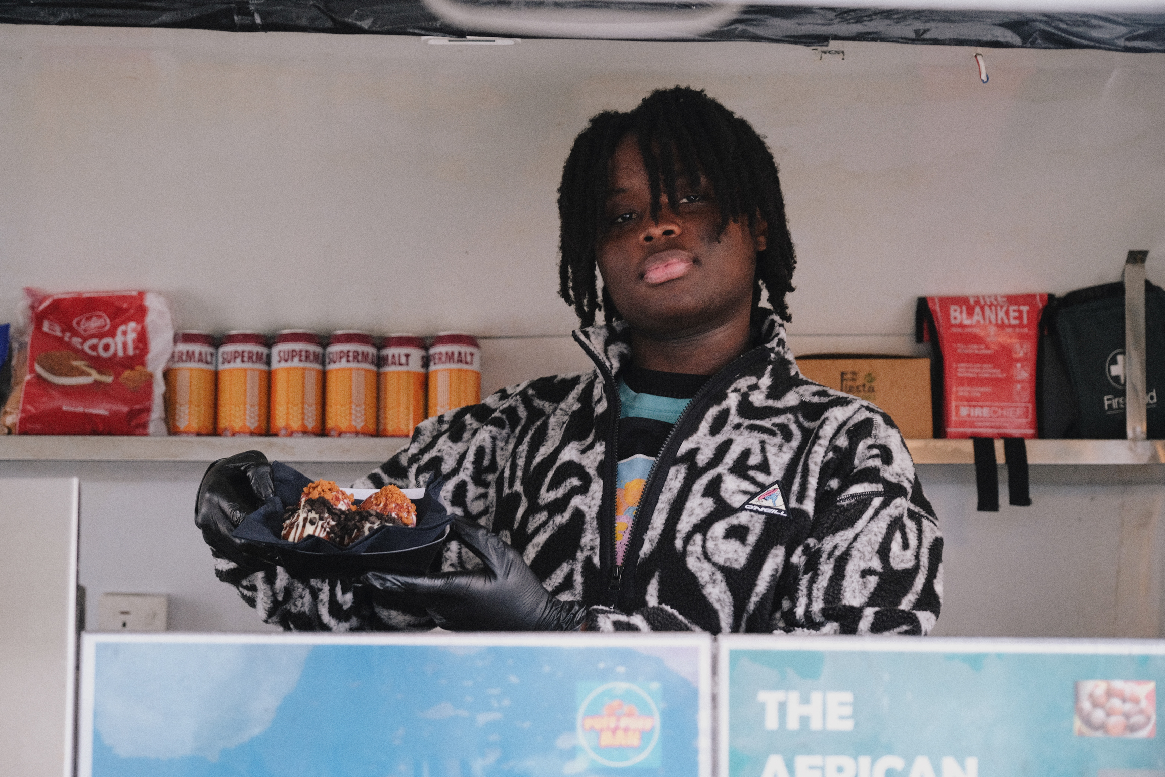 Person with dreadlocks wearing black and white patterned jacket behind shop counter with shelves of products and drinks