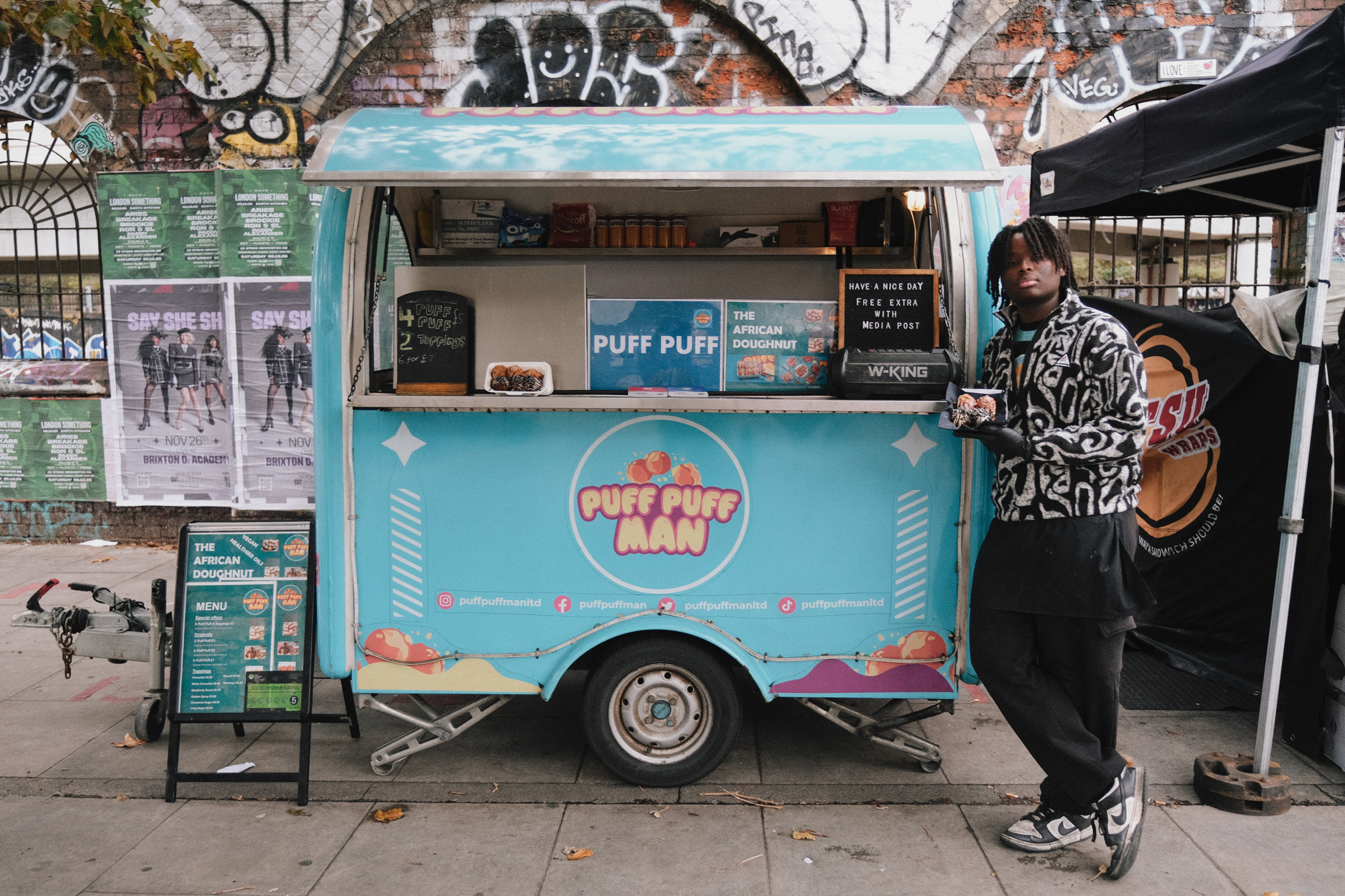 Turquoise food cart with colourful graphics and serving window, person in patterned coat standing beside it against graffitied brick wall.