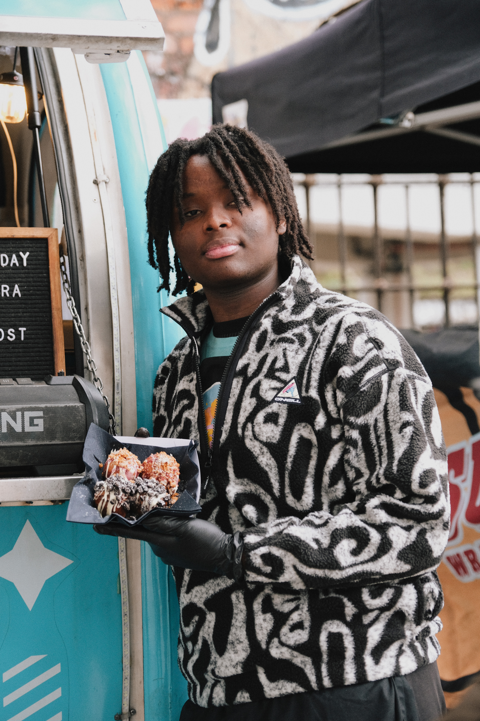 Young person with dreadlocks wearing patterned black and white jumper holds food item whilst standing beside blue food truck.