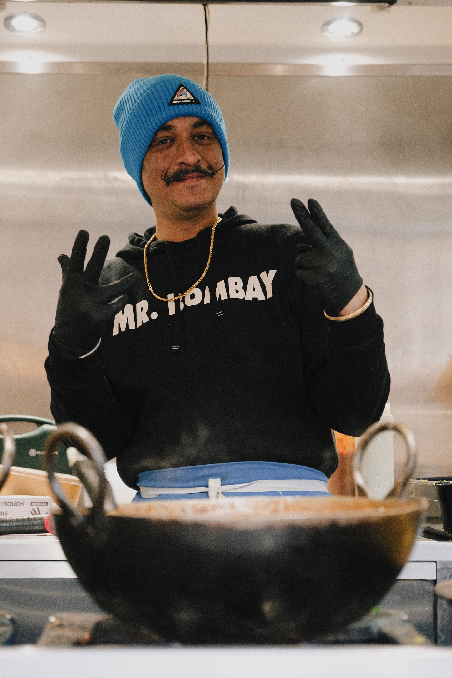 Man in blue beanie and black "MR. BOMBAY" jumper making peace signs with black gloves in kitchen with large metal bowl.