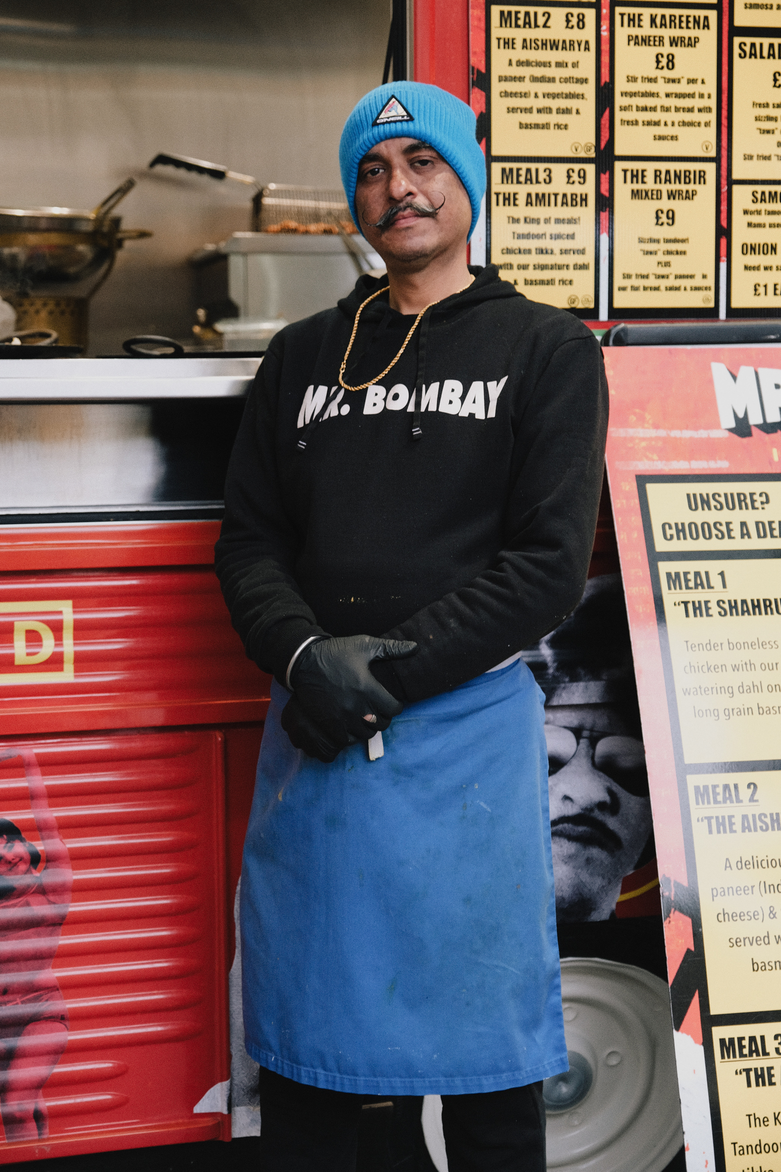 Man in blue beanie and apron wearing black "Mr. Bombay" hoodie stands in takeaway kitchen with menu boards and red shutters visible.