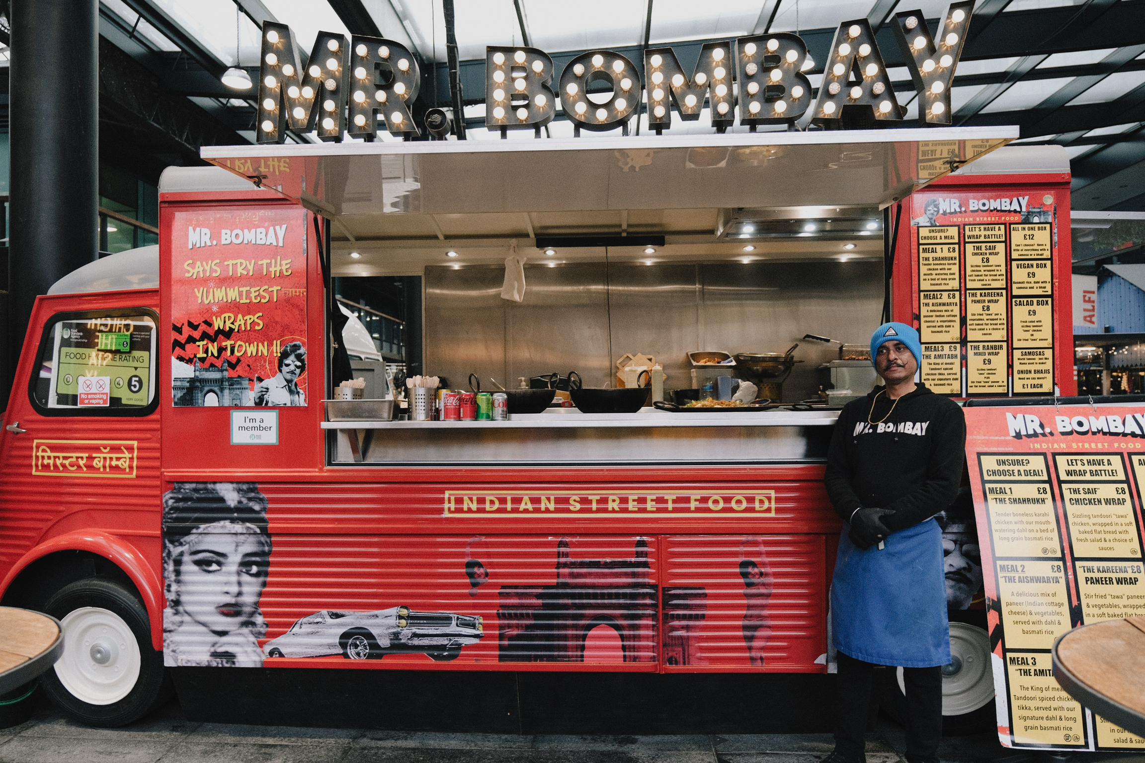 Red food truck with "MR. BOMBAY" lettering in lights above. Staff member in blue apron stands beside truck with menu boards visible.